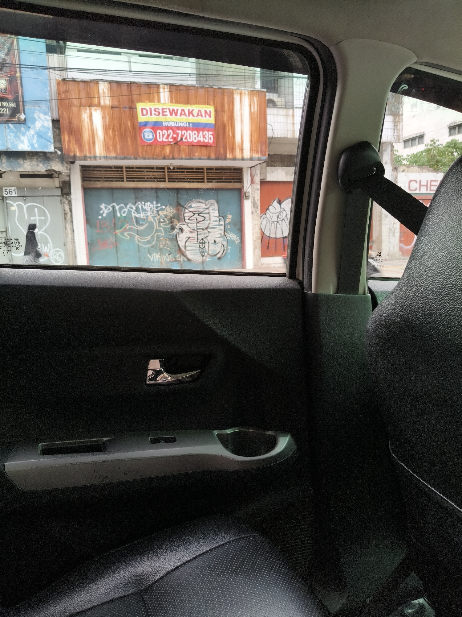 A sign reading "DISEWAKAN" (for rent) with the contact number "022-7208435" is prominently displayed on a rusted corrugated metal storefront. The storefront is directly across from the interior of a car, viewed through the rear window. To the left of the storefront, a blue metal door is covered in graffiti, including a depiction of a stylized character that resembles a tiger. A person wearing a dark jacket and a face mask walks past the blue door. The number "561" is visible on a light blue surface above the door. To the right of the storefront, a white building with windows is visible, and the word "CHE" is painted on its exterior. The scene appears to be a street in Bandung City, Indonesia, viewed from inside a vehicle. The car's interior shows a black leather seat, a silver door handle, and a cup holder.