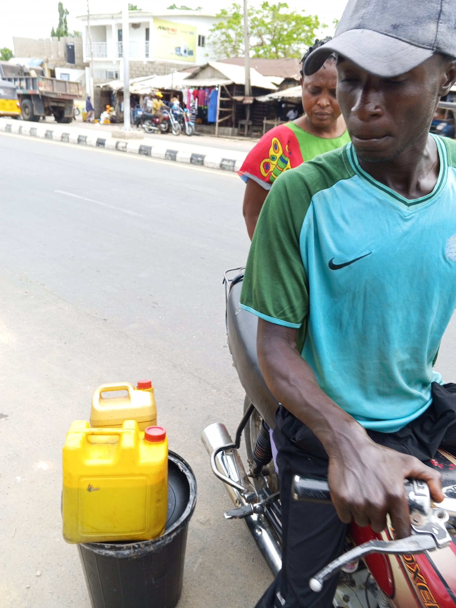 The image depicts a man in a blue and green sports shirt and a gray cap sitting on a motorcycle with a woman behind him on a street in Zango-ndaji, Nigeria. Two yellow gas canisters rest in a black bucket near the motorcycle. Behind them is a street with buildings and a few people visible in the background.