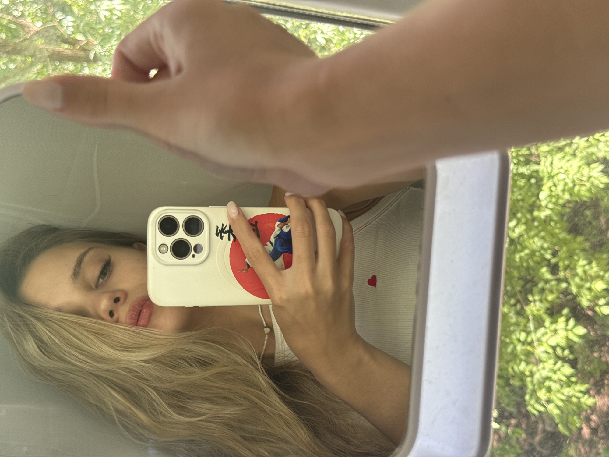 The image depicts a person taking a selfie in a car mirror. The person has long, blonde hair and is wearing a white tank top with a small red heart design. They are holding a smartphone with a white case. The phone case has a red circle with Japanese characters and an image of two figures. The reflection shows a partial view of the person's face, including their eye, nose, and mouth. The mirror is bordered by white trim. Through the car window, green foliage is visible. The location of Chaikovsky, Russia cannot be determined from the image.