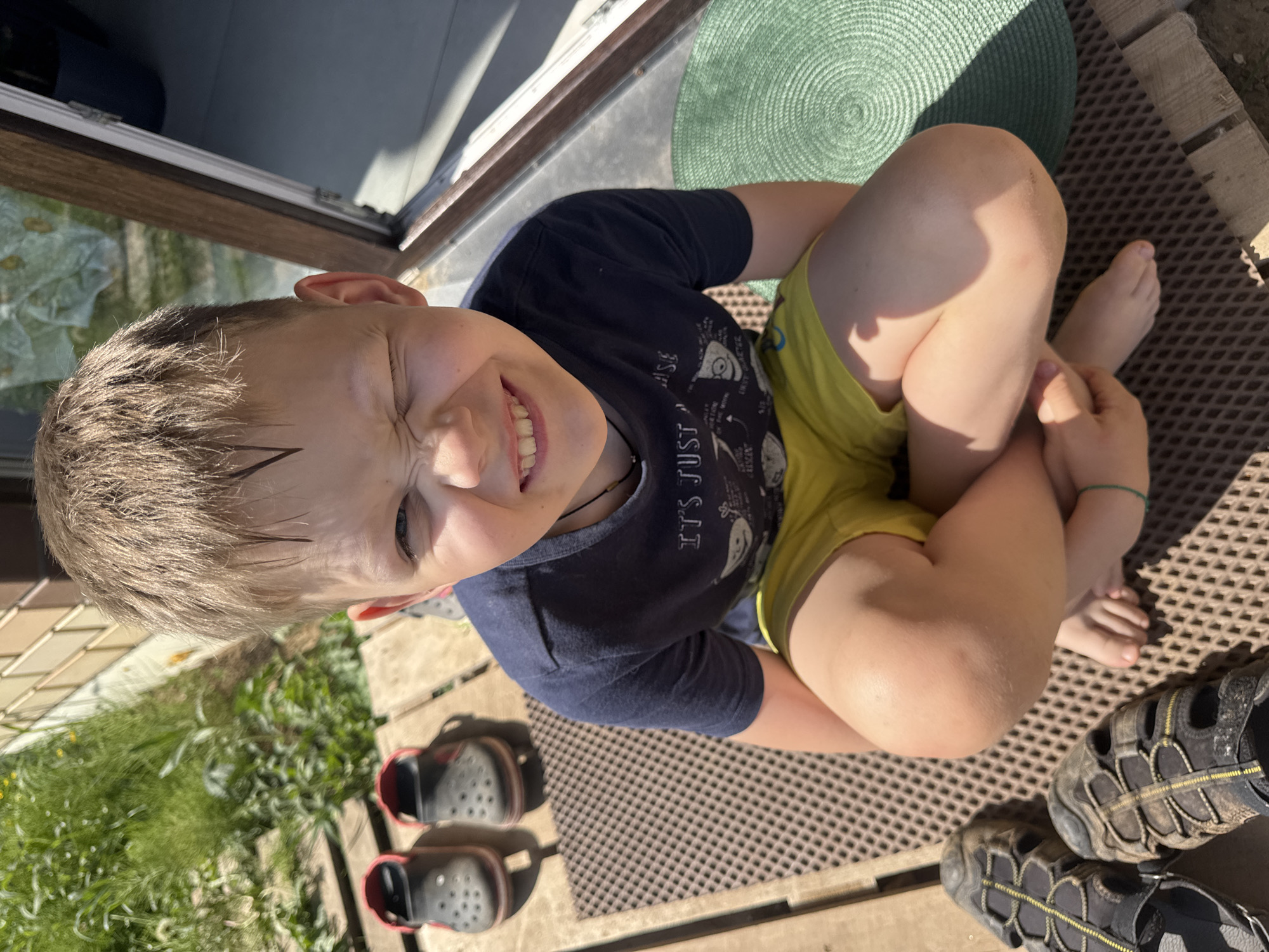 A boy with blond hair is sitting cross-legged on a brown, textured outdoor mat. He wears a navy blue shirt with graphics and yellow shorts. He is smiling and squinting slightly. There are a pair of red and black Crocs and a pair of dirty, multi-toned sandals on the mat near him. To his right is a round green mat. There is some foliage and a section of a wooden door frame visible. The scene is brightly lit, indicating an outdoor location. The location is identified as Ферма, Russia.