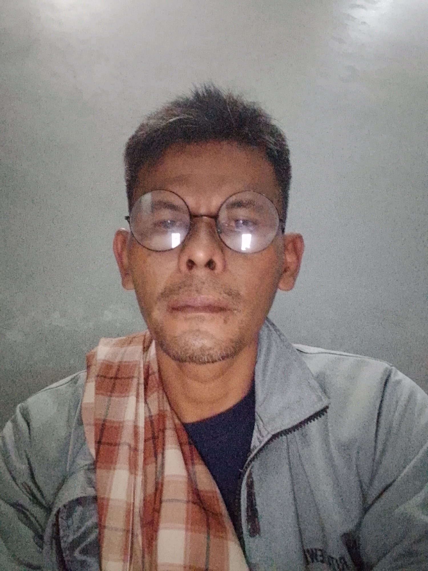 A man with short, spiky dark hair and a light stubble is pictured from the chest up, looking directly at the camera. He is wearing round, thin-rimmed glasses. Around his neck, he has a checkered scarf in shades of orange, brown, and white. He is also wearing a light gray zip-up jacket over a dark blue shirt. The background is a plain, textured gray wall. The image appears to have been taken indoors, likely in a residential setting, given the simple background and the casual attire of the subject. There is no clear indication of the specific location within Padang Dalom, Indonesia, beyond the fact that it is a person's portrait.