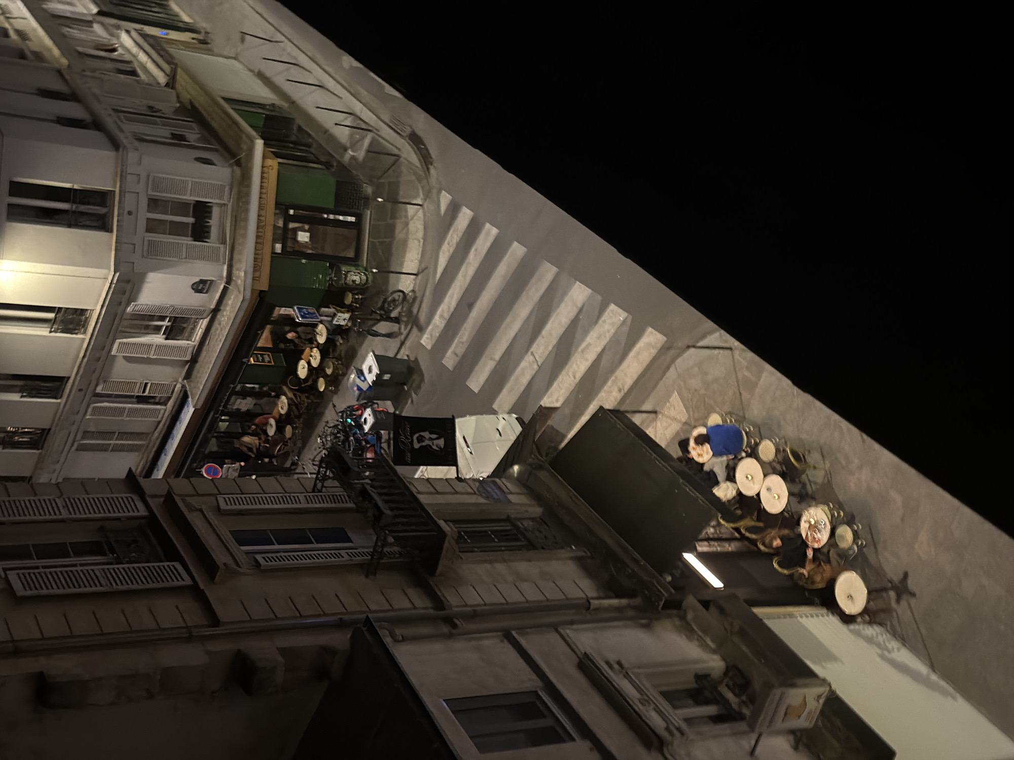 The image, rotated 90 degrees counter-clockwise, captures a nocturnal street scene in what is identified as Paris, France. On the left side of the frame, several multi-story buildings with classic Parisian architecture line the street. These buildings feature numerous windows, many adorned with closed white or dark shutters. Some lower-level units appear to be commercial establishments, one notably sporting bright green awnings and another exhibiting outdoor seating.
The center and right portions of the image display the street itself, marked by a clear pedestrian crosswalk with white stripes. Adjacent to the crosswalk, a significant area is arranged with numerous small, round tables and chairs, typical of an outdoor cafe or restaurant terrace. These tables and chairs mostly appear unoccupied and some are stacked, suggesting the late hour or closure. A single bicycle is parked nearby. The upper right corner of the image is completely dark, confirming the nighttime setting.
Artificial lighting, likely from streetlights and interior building lights, illuminates parts of the scene, creating strong contrasts and shadows, particularly from the buildings onto the street. The overall ambiance is quiet and deserted.
Visible text includes:
- On an awning above a shop with green awnings: "DU VIEUX MARCHE DES M..." (partially obscured).
- On a black flag or banner hanging near the outdoor seating: A white graphic (possibly a stylized face or logo) and some white text, where "Paris" might be partially visible.
- A street sign on one of the buildings reads "RUE..." (partially obscured).
No people are visible in the scene, and no specific activity is currently taking place. The weather appears clear.