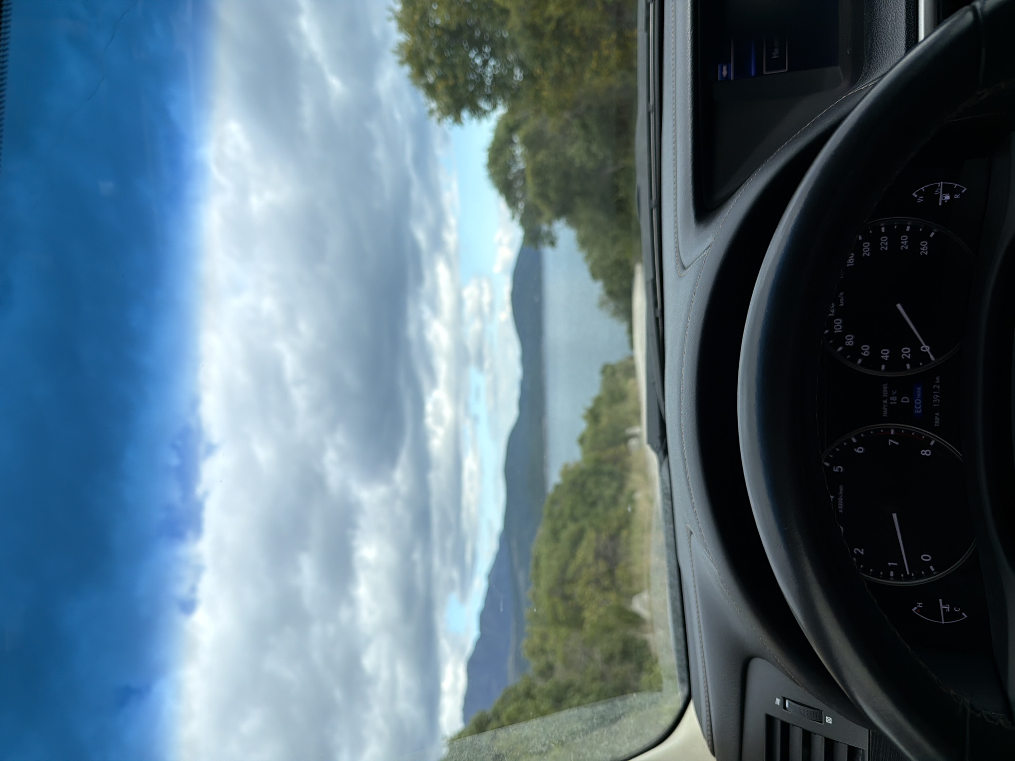 A view from inside a car shows a winding road with mountains and a lake in the distance. The speedometer indicates the car is not moving. The digital display shows "TRIPA 1391.2km", suggesting the car has traveled this distance. The dashboard also shows the temperature is 18°C, and the car is in drive. The background includes a landscape of trees and a lake, partially obscured by clouds. While the city cannot be confirmed with absolute certainty, given the landscape and proximity to the lake, Radovići, Montenegro seems like a possible location.