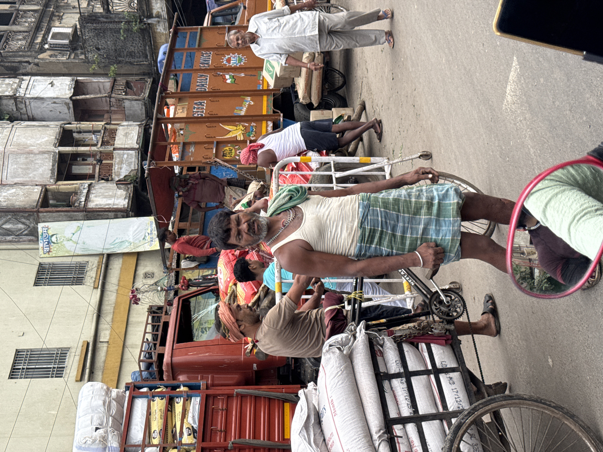 Here is a detailed description of the image:
**Location:** The image appears to be taken on a street in Kolkata, India. The presence of trucks with Bengali writing and the type of rickshaws suggest this location.
**People:** There are several men in the image. Some appear to be manual laborers pulling or pushing rickshaws loaded with goods. One man is wearing a white shirt and light-colored pants, seemingly supervising the work. The men wear clothing typical of manual laborers, such as sleeveless undershirts and turbans.
**Subjects:** The main subjects are the cycle rickshaws loaded with sacks of goods, likely being transported for commercial purposes. A red truck is also visible and filled with goods.
**Objects:** Key objects in the image include:
* Cycle rickshaws
* Sacks filled with goods
* Trucks loaded with materials
* Buildings in a state of disrepair
* A mirror reflecting the surroundings.
**Actions and Interactions:** The primary action depicted is the transportation of goods using manual labor (rickshaws). There are interactions between the laborers and a person who appears to be overseeing the operation.
**Environment:** The environment is characterized by a busy street with buildings in the background that show signs of age and disrepair. The image suggests a commercial area with transportation activities.