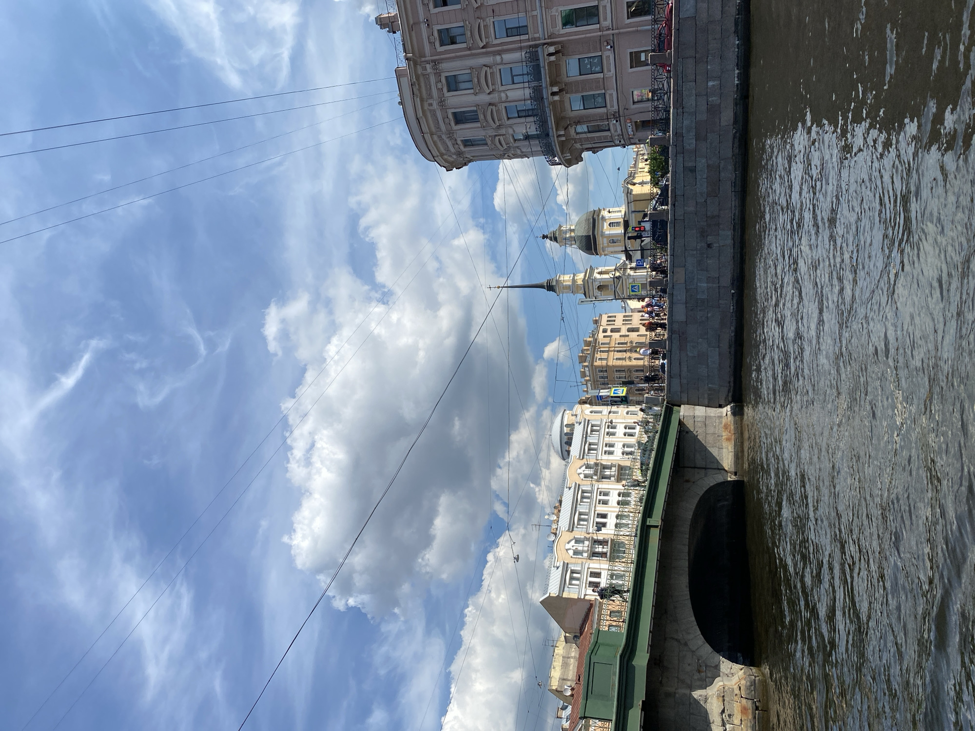 This image shows a view of a canal in Saint Petersburg, Russia, from the water level. On the left, a stone bridge with a green railing arches over the canal, with a dark opening beneath. Beyond the bridge, several buildings with ornate facades line the canal bank. In the middle ground, a yellow building with a tall spire and a dome is visible, suggesting it is a church or religious structure. To the right, a multi-story building with a curved facade and balconies stands prominently. The sky above is blue with scattered white clouds, and several power lines stretch across the frame. People are visible walking on the bridge and the embankment. The water in the canal is dark and reflects the light.
