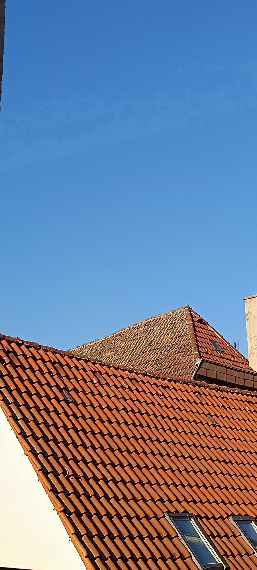This image shows a close-up of two adjacent tiled roofs against a clear, bright blue sky. The roof in the foreground is covered in vibrant, orange-red tiles that are laid in a regular pattern. Small, curved metal hooks are visible along the eaves of this roof. Behind it, a second roof is visible, with tiles that appear older and more weathered, displaying a mix of brown and orange hues. This upper roof has a steeper pitch and is partially obscured by the foreground roof. A small skylight is visible on the upper roof, along with a darker section of trim. To the right of the upper roof, a portion of a beige, possibly stucco, wall or structure is visible. The overall scene suggests an outdoor setting, likely during the daytime given the bright sunlight casting shadows on the tiles, indicating a clear day. The tiled roofs and the style of the building suggest a European location, consistent with Winnenden, Germany. There are no people or animals visible in the image. The focus is entirely on the architectural details of the rooftops and the expanse of the sky.