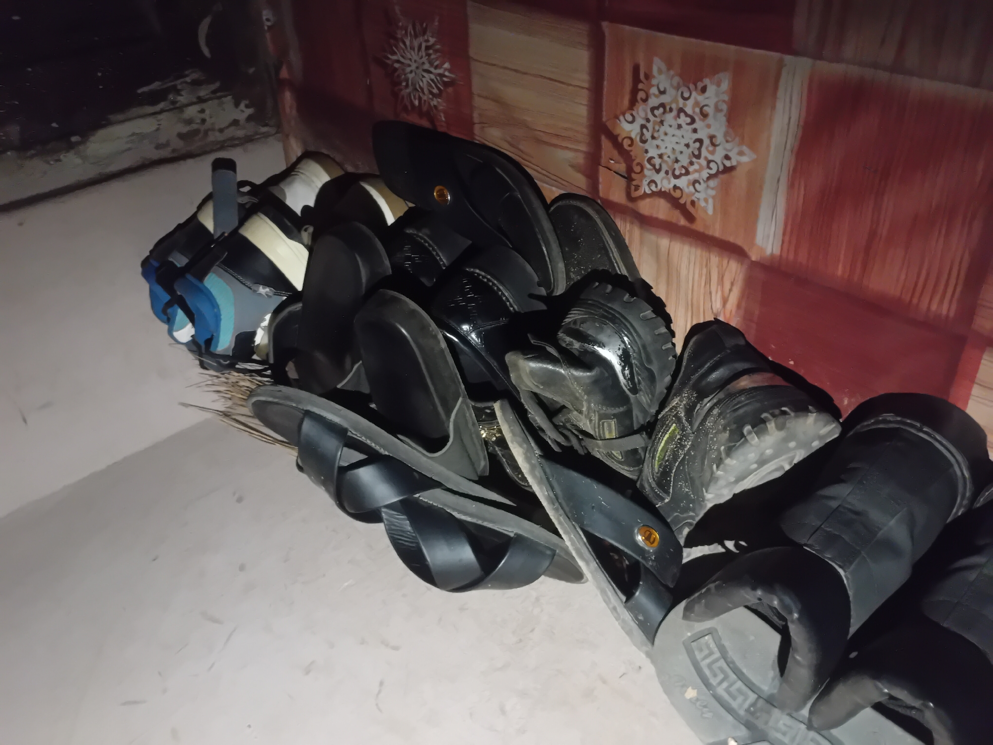 A pile of footwear is stacked against a light-colored wall, with a patterned floor visible to the right. The footwear includes several pairs of black sandals, including a cross-strap style, and a pair of black patent leather shoes. At the top of the pile, a pair of multi-colored sneakers, featuring blue, black, and white accents, is visible. The floor has a repeating pattern of reddish-brown and tan stripes, with decorative snowflake-like designs. The scene is dimly lit, suggesting an indoor environment, possibly a storage area or entryway in Jauro Yinu, Nigeria.