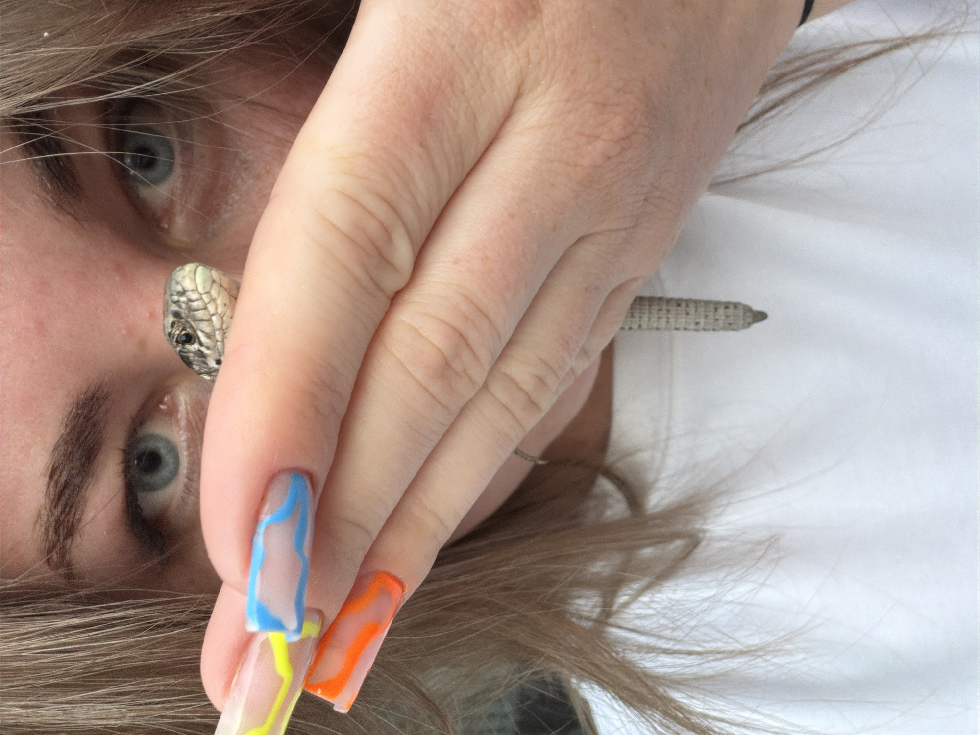 A close-up, rotated portrait shot features a person with light hair and blue eyes, partially obscured by a hand. The person's left eye and eyebrow are visible, gazing directly forward. Light brown or blonde hair frames the face and spreads across the frame.
A hand with long, clear acrylic nails, adorned with vibrant wavy designs in blue, yellow, and orange, is positioned across the person's face. The designs appear to be painted on top of the clear base. Resting near the person's nose and eye is a metallic, textured object resembling a snake's head, complete with scales and a small black eye, possibly a ring or a piece of jewelry. In the background, to the right of the hand, another slender, textured metallic object, resembling a pen or stylus, is faintly visible against a plain white background. The lighting is bright and even, suggesting an indoor setting or diffused daylight. No discernible text is present. The overall image presents a stylized and artistic composition, focusing on intricate details like the nails and the snake-like accessory.