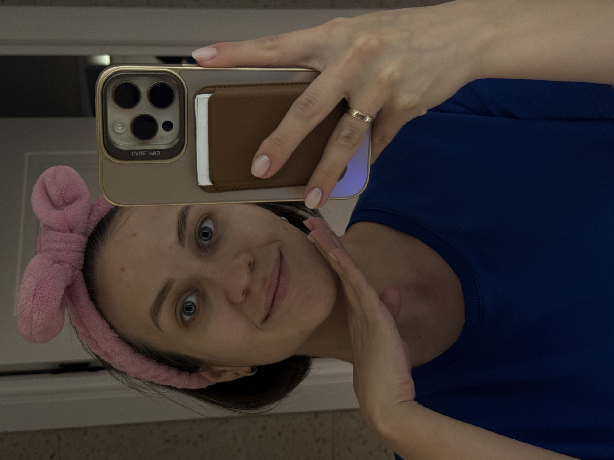A young woman is taking a selfie. She's wearing a blue shirt and a pink headband with a bow. She's holding a smartphone with a brown wallet-style case that has slots for cards. She has a ring on her finger. The background is blurry, but it appears to be a bathroom with light-colored walls. No information to suggest a location in the city of Pervomaysky, Russia.