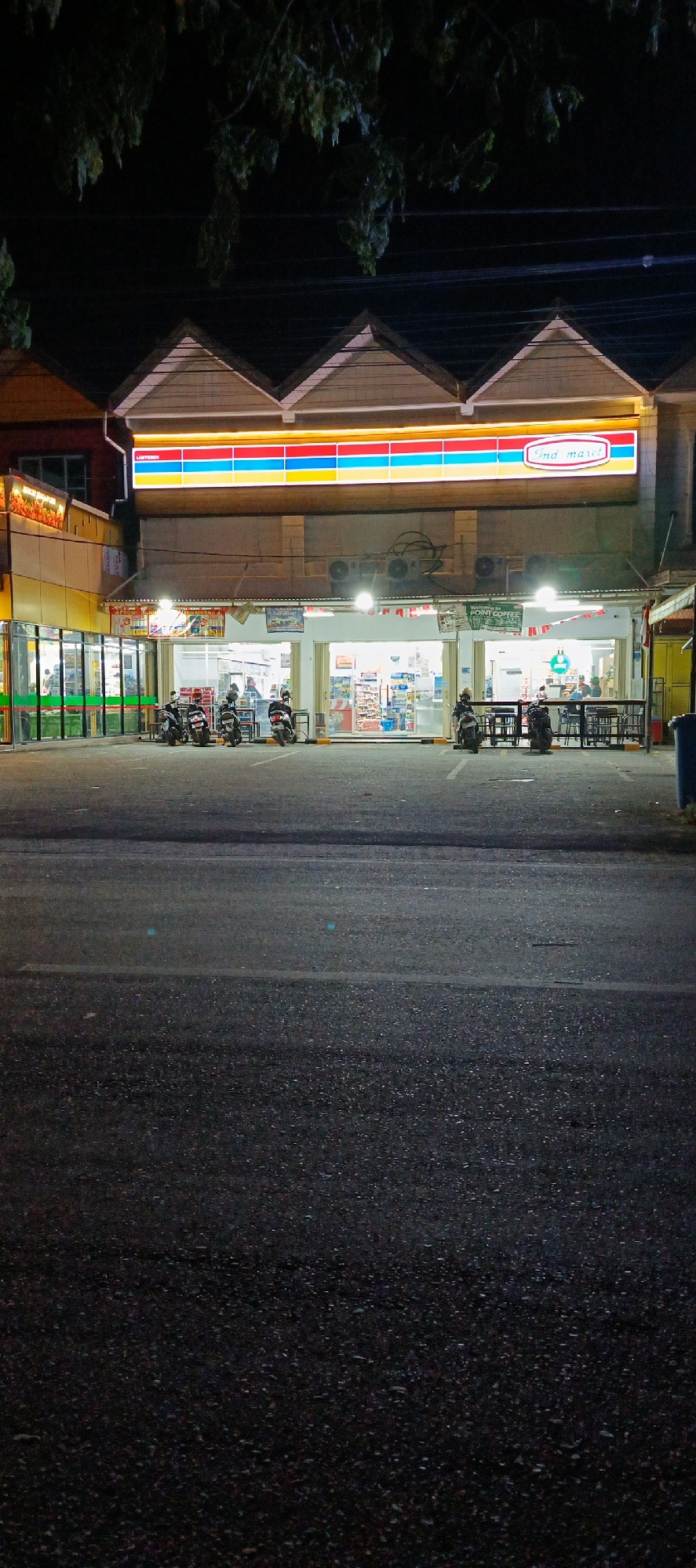 The image depicts a nighttime scene in front of an Indomaret convenience store located on Jalan Teuku Umar in Banda Aceh, Indonesia. The store's facade is brightly lit, with a prominent sign featuring horizontal stripes of yellow, red, blue, and white, and the Indomaret logo in the upper right corner. Multiple motorcycles are parked in front of the store, and several people are visible inside, browsing shelves and interacting with staff. The area in front of the store appears to be a parking lot, with asphalt paving and visible parking lines. The surrounding environment suggests a commercial street with other buildings partially visible. The overall atmosphere is one of a typical urban commercial hub operating during evening hours.