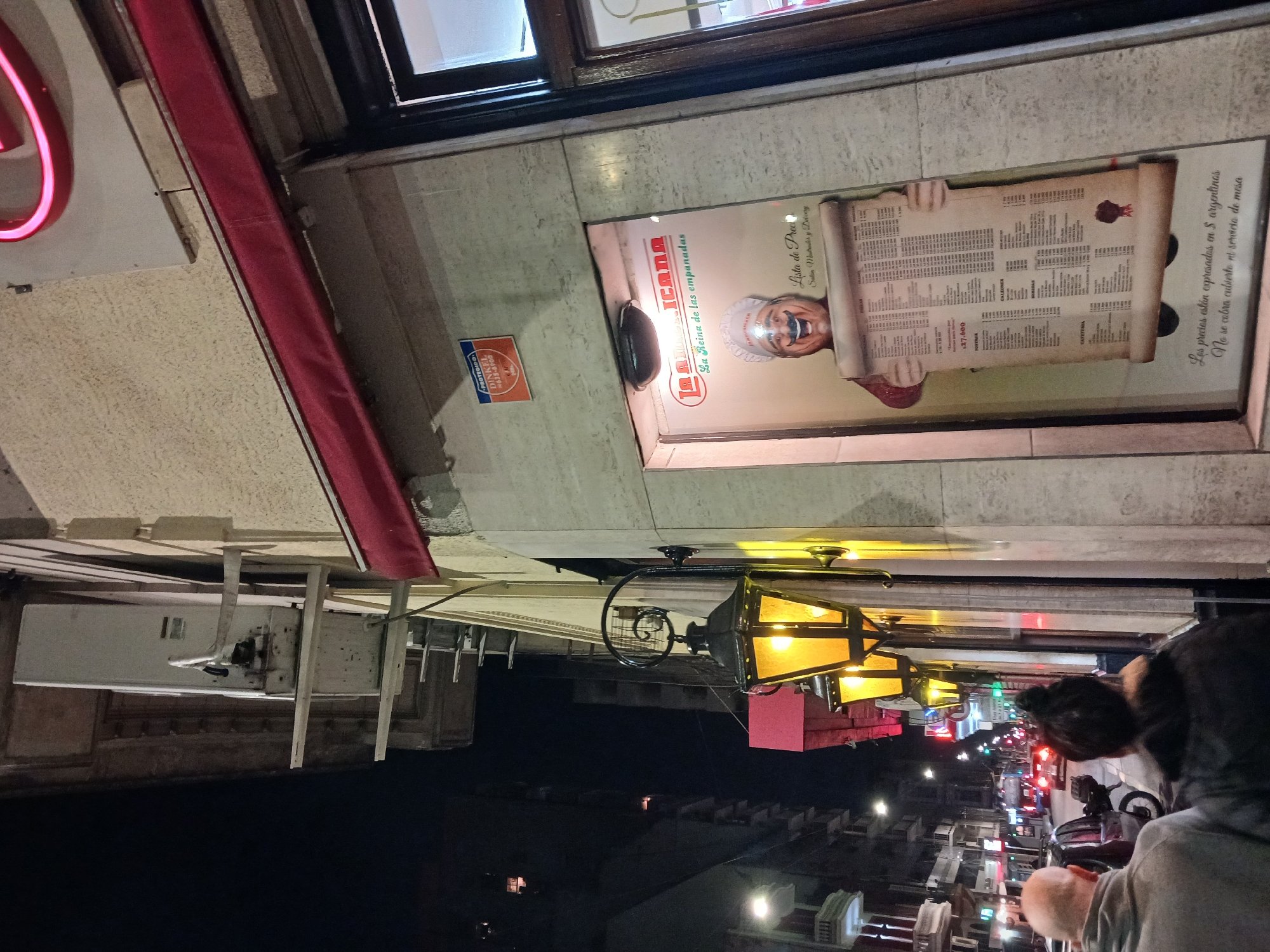 The image shows a street in Buenos Aires, Argentina, at night. The camera is pointed upward toward the front of a business, "La Reina de las empanadas" (The Queen of Empanadas). A menu is displayed with the name of the shop and the phrase, "Lista de Prea" (Price List), with details about the cost of food and coffee. A sign at the bottom reads, "Los precios están expresados en $ argentinos" (The prices are expressed in Argentine pesos). Street lights are visible down the street, and a few people can be seen.
