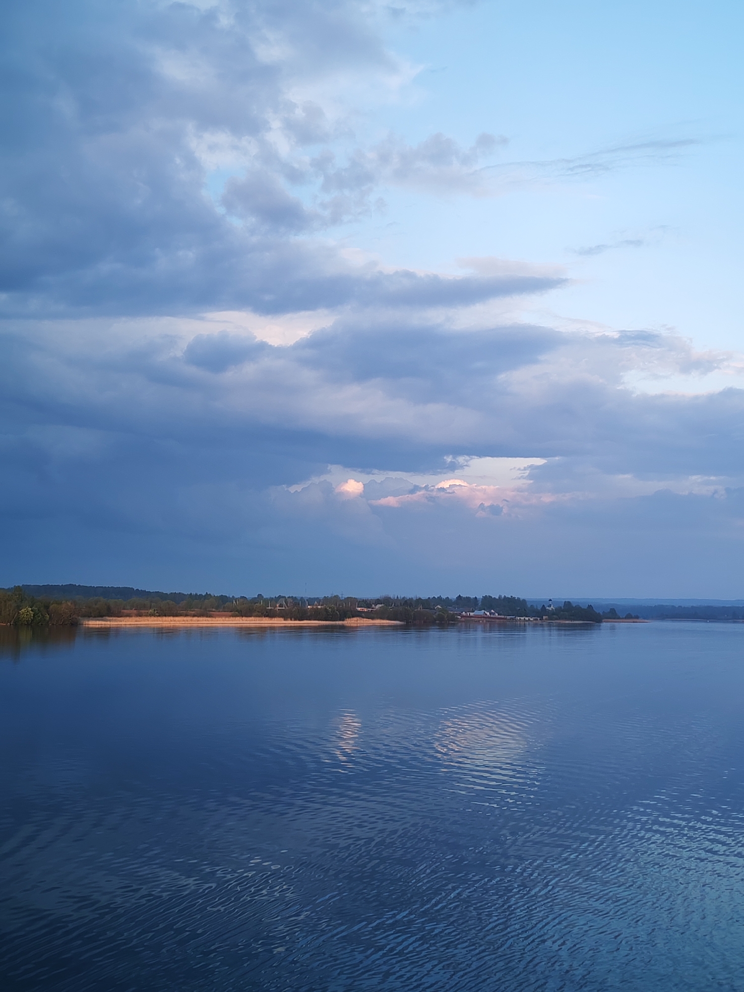 The image depicts a tranquil lake scene under a cloudy sky. A body of water fills the majority of the frame, reflecting the light and clouds. A shoreline with trees and vegetation runs horizontally across the middle distance, with buildings and a church steeple visible on the shoreline. The sky is filled with gray and white clouds, with some hints of blue sky peeking through. The light suggests either early morning or late evening.
The image location can be identified as Ankhimovo, Russia, based on the presence of a church with a distinct dome. The shoreline and water match the setting of Lake Beloye, which is located near Ankhimovo.