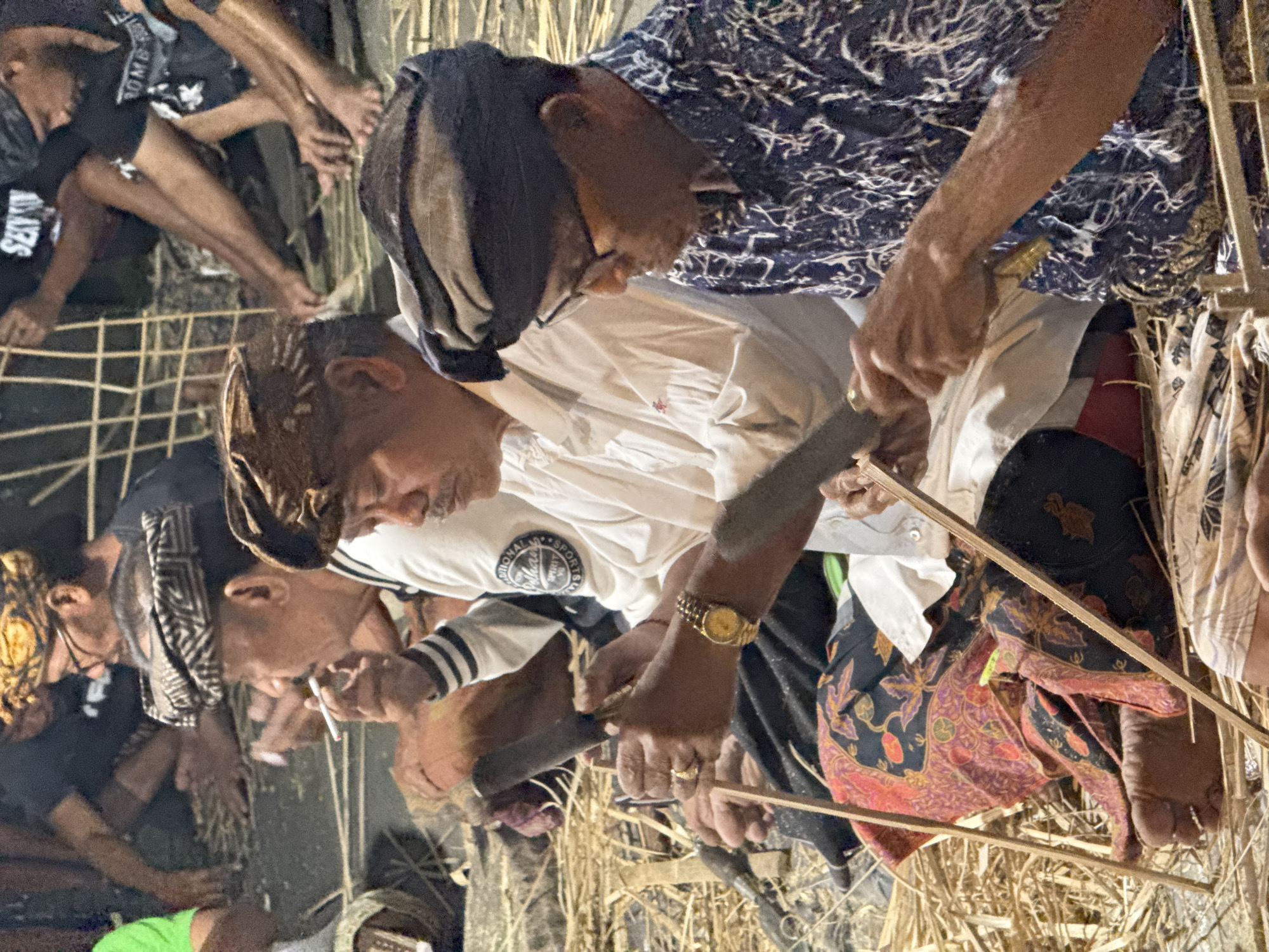 Three men are seated on the ground, surrounded by dried straw and bamboo poles. The man in the center is wearing a white long-sleeved shirt and a brown headscarf, and is smoking a cigarette. He is holding a wooden tool in his right hand, as if preparing to carve or work on something. To his left, another man in a black and white striped shirt and a patterned headscarf is smoking. He holds a cigarette in his mouth and appears to be focused on an unseen task. To the right of the central figure, a third man wearing a dark blue patterned shirt and a black headscarf is using a knife to work on a bamboo pole. He is also wearing glasses. The men are dressed in traditional attire, suggesting they might be participating in a local ceremony or craft activity. The environment appears to be outdoors, possibly in a communal gathering space in the Ubud District of Indonesia, given the distinctive cultural context suggested by their clothing and the materials present.