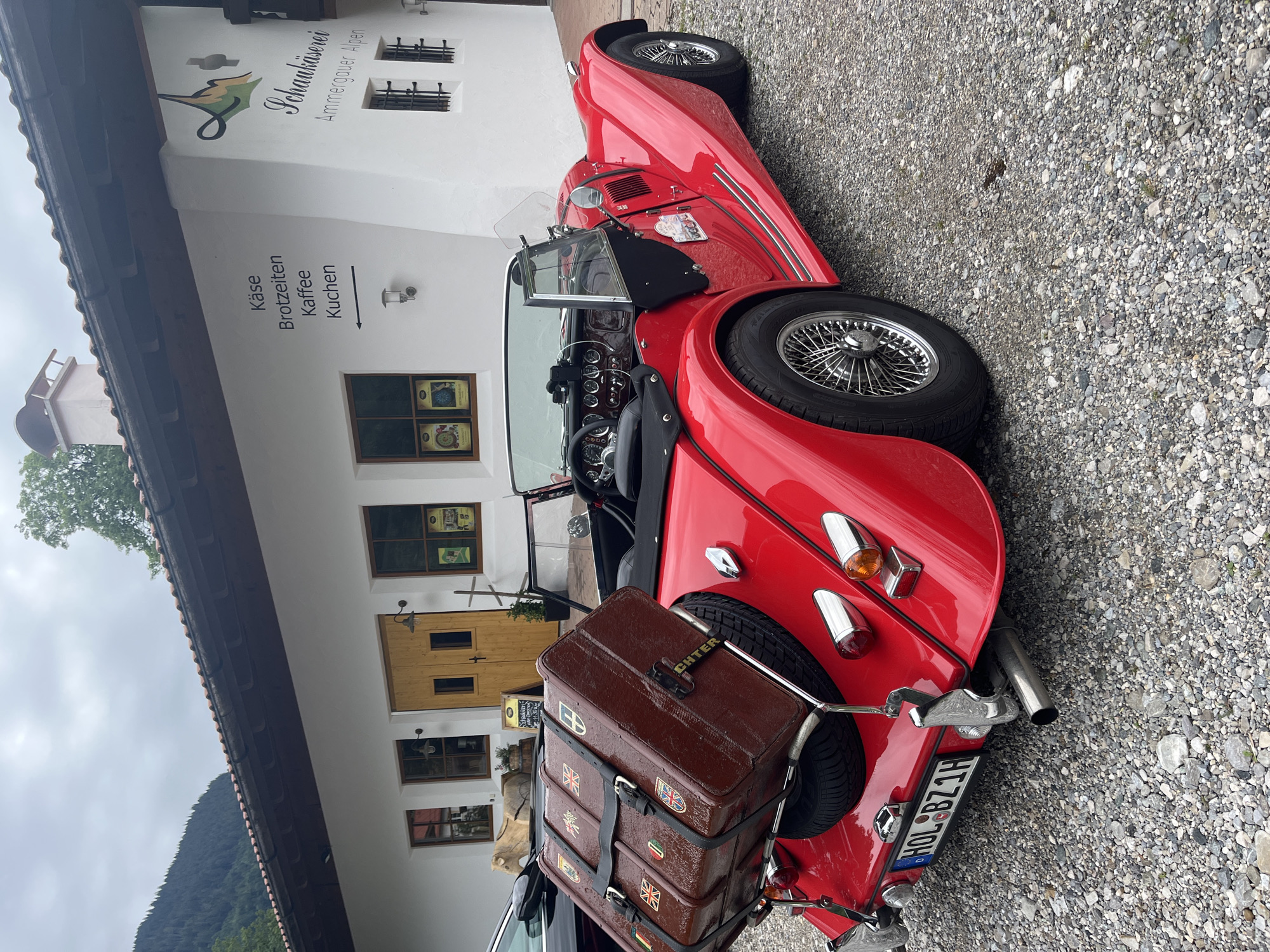 A bright red convertible sports car is parked in front of a white building in Ettal, Germany. The car has its trunk open, revealing a spare tire and a large, brown vintage-style suitcase secured on top. The suitcase is adorned with various travel stickers from different countries. The building appears to be a shop or restaurant, with signage indicating "Käse," "Brotzeiten," "Kaffee," and "Kuchen," which translate to "Cheese," "Snacks," "Coffee," and "Cake." Additional signage above the door reads "Schaukäserei Ammergauer Alpen," meaning "Show Dairy Ammergau Alps." The building has traditional wooden doors and windows. The ground is covered in gravel. In the background, a lush green mountain slope is visible under a cloudy sky.