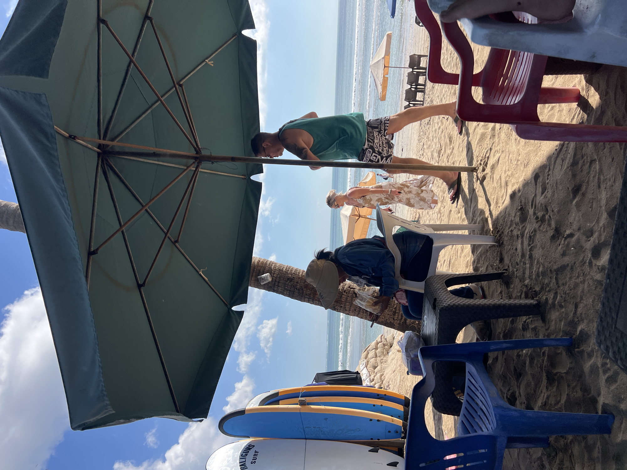 The image depicts a beach scene in Kuta, Indonesia, likely a tourist area given the surfboards. In the foreground, a person wearing a wide-brimmed hat and a denim jacket sits at a small black plastic table on the sand, holding a food item. To their left, a blue plastic chair is visible. Behind them, several surfboards are propped up against a low wall or structure. A large green umbrella provides shade, supported by a pole that extends upwards.
In the mid-ground, a man in a green tank top and patterned shorts walks past the umbrella, while a woman in a floral dress walks behind him. The ocean stretches out in the background, with gentle waves and a clear blue sky with scattered clouds. Additional beach umbrellas and chairs are visible further down the beach. A palm tree trunk is present near the seated person, partially obscured by the umbrella. The sand appears dark in the foreground, suggesting it may be wet or mixed with volcanic ash.