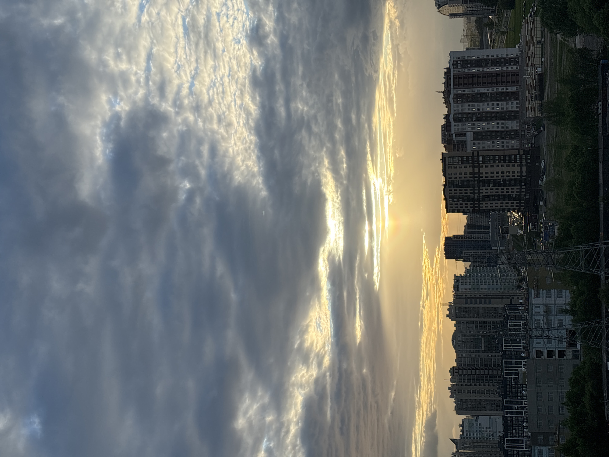 The image depicts an urban environment under a dramatic sky. The setting sun casts a golden light on the clouds and the buildings below. The buildings are high-rise residential structures with multiple windows. A dense cloud cover fills the left side of the frame, while the sun appears to be setting on the right, creating a contrast between light and shadow. Power lines are also visible, adding to the urban scene.
I am unable to provide the exact location in the city of Unknown, Unknown, as I do not have access to such location data.