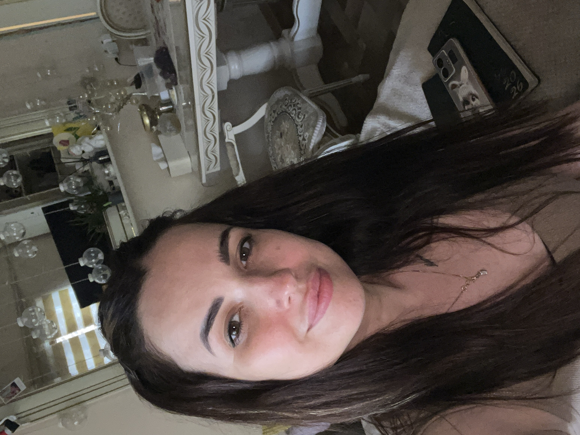 A woman with long, dark hair and brown eyes is taking a selfie indoors. She is wearing a light-colored top and a necklace. In the background, a dining table with a white tablecloth and ornate trim is visible, along with a chair. A mirror reflects blinds and decorative lights. A phone and a notebook are on a surface in the foreground.