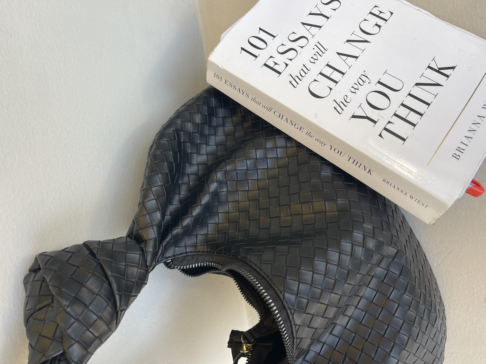 A black woven handbag is positioned diagonally across the frame, partially obscuring a white book. The handbag is made of a textured material with a braided pattern and features a silver zipper. The book is lying on a white surface, with its cover displaying bold black text that reads "101 ESSAYS that will CHANGE the way YOU THINK". The author's name, "BRIANNA WIEST", is printed in smaller text on the spine. A red object, possibly a pen, is partially visible sticking out from the top of the book. The background is a plain, textured white surface. The image appears to be a close-up still life, with no people or identifiable landmarks of North Kuta, Indonesia present.