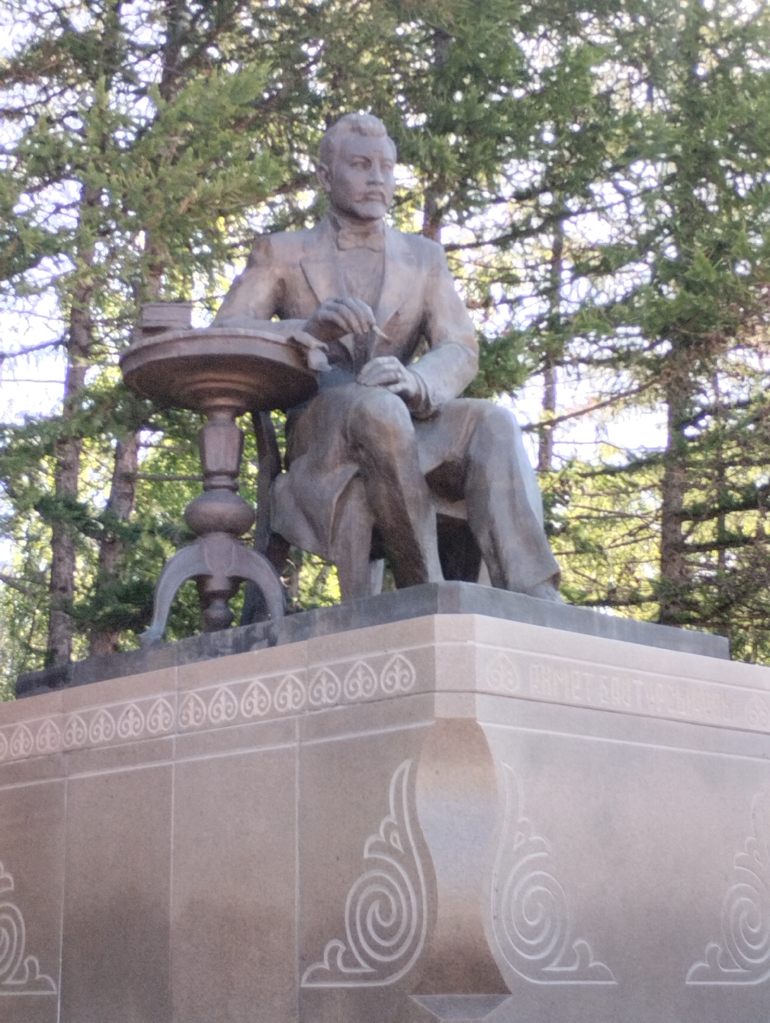 A bronze statue depicts a seated man in formal attire, including a jacket, trousers, and a bow tie. He is holding a pen in his right hand and appears to be writing on a book or document placed on a small, round table beside him. The statue is mounted on a granite pedestal with decorative carvings. In the background, tall trees with dense foliage are visible, suggesting an outdoor setting. The image is taken from a low angle, looking up at the statue and pedestal. The text inscribed on the pedestal appears to be in Cyrillic or Latin script and is partially obscured by the angle and lighting. This monument is located in Kostanay, Kazakhstan, and is dedicated to the writer and poet Akhmet Baitursynov.