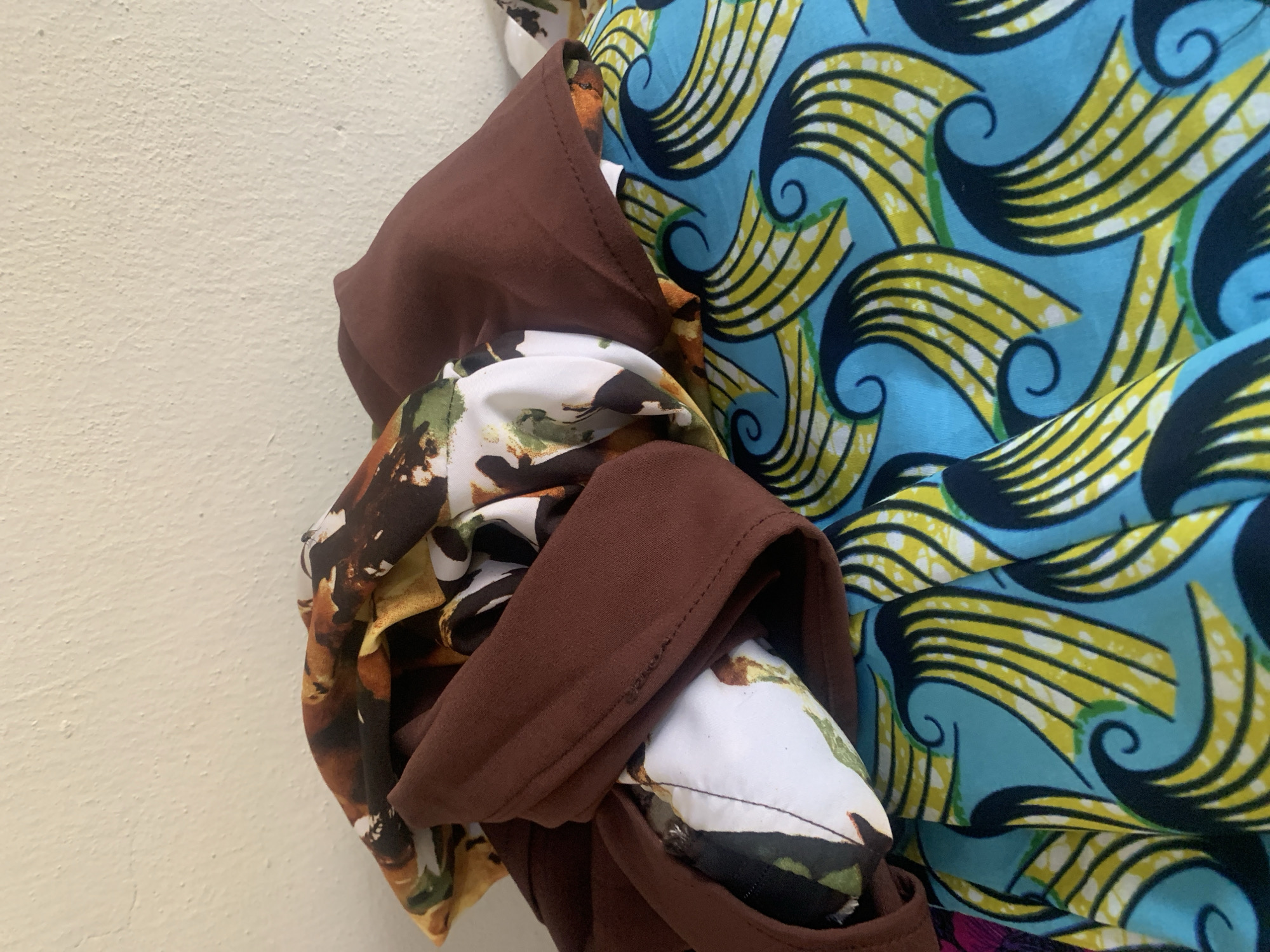 A close-up shot shows a pile of fabric with a cream-colored wall in the background. The fabric has a variety of patterns, including a brown fabric folded over a white fabric with a floral print. Below that, a bright blue fabric with a yellow and black wave-like pattern is visible. The scene appears to be indoors, possibly in a home or workshop. There is no indication of the city of Jauro Yinu, Nigeria, in the image.