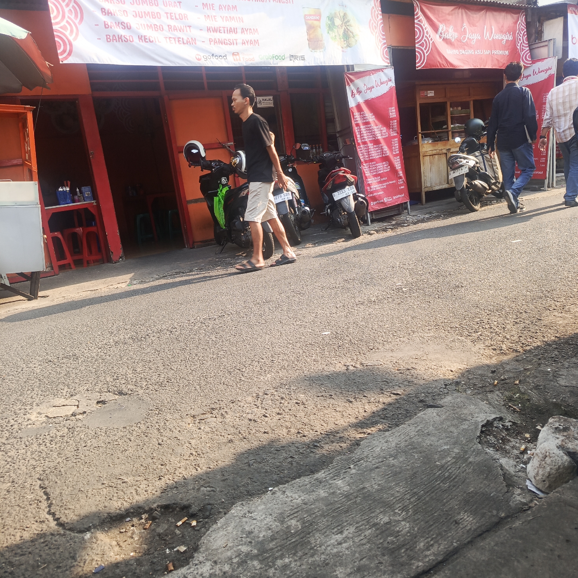 In South Jakarta, Indonesia, a street scene unfolds in front of a food establishment, likely a "bakso" (meatball) stall, with its name "Bakso Jaya Wonogiri" prominently displayed on red banners. The establishment features an orange facade with wooden counters and glass windows. Various food items and prices are listed on the banners, indicating offerings like "Bakso Jumbo Urat," "Mie Ayam," and "Kwewtiaw Ayam."
Several motorcycles are parked on the roadside, suggesting a common mode of transportation in the area. A man, dressed in a black t-shirt and light-colored shorts, walks past the motorcycles, his posture suggesting he is heading away from the establishment. Further down the street, two men, one in a dark shirt and jeans, and another in a checkered shirt and jeans, are walking away from the camera, towards the right side of the frame.
The street itself appears to be paved with asphalt and shows signs of wear and tear, with visible cracks and uneven surfaces. The presence of cigarette butts scattered on the road and sidewalk indicates recent activity. The environment is illuminated by natural sunlight, casting shadows from the buildings and objects onto the street. The overall impression is that of a typical urban street scene in Indonesia, characterized by local businesses, everyday life, and a busy atmosphere.