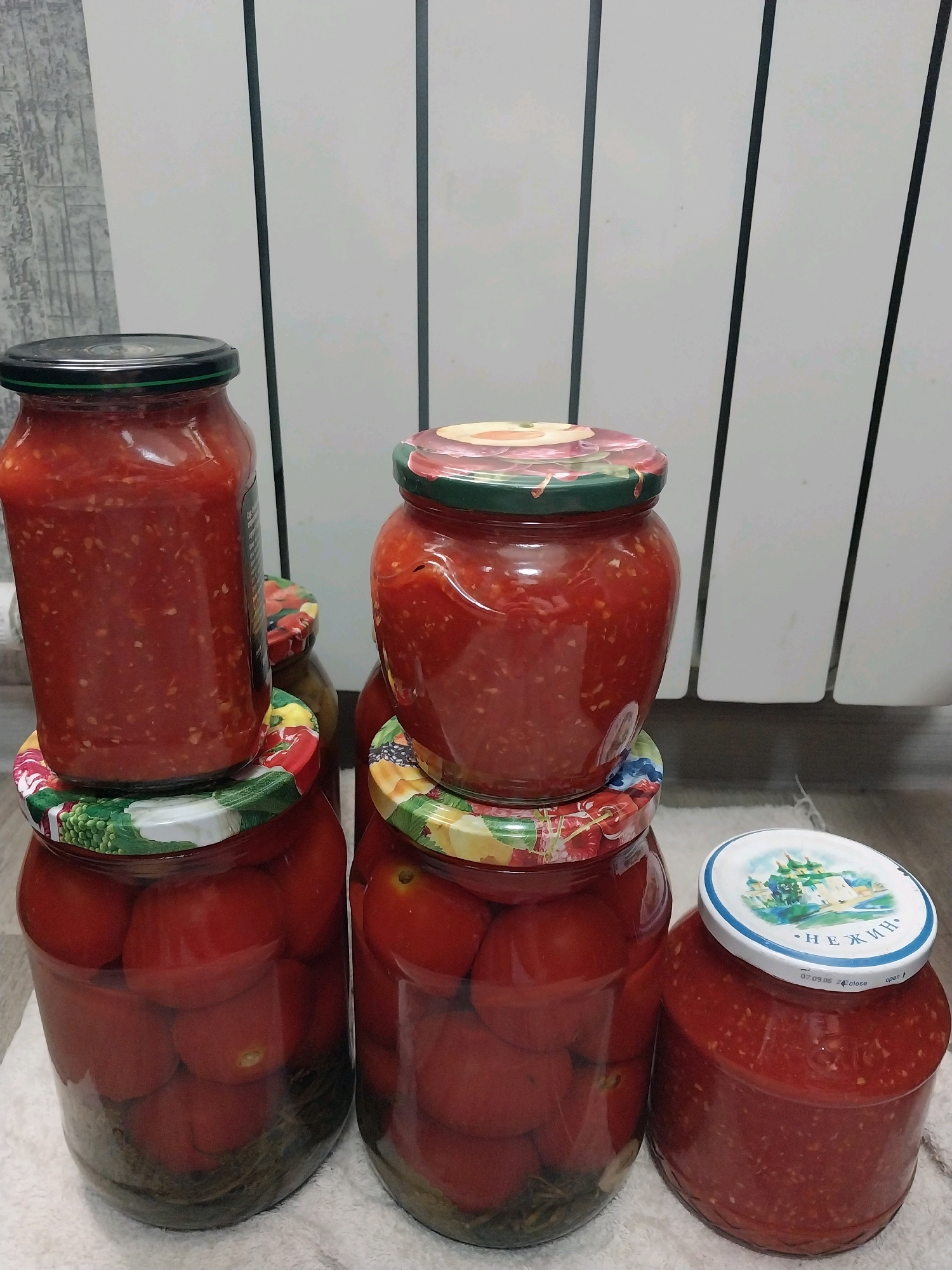 The image shows a collection of six glass jars filled with red food items, likely tomatoes and tomato-based products. The jars are placed on a white cloth in front of a white radiator with vertical slats. The jars are of various sizes and shapes, with several having decorative lids. Some jars contain whole tomatoes submerged in liquid, while others are filled with a chopped or pureed red substance. The lighting is dim, and the background is out of focus. There are no people or animals visible in the image, and no specific location within the city of Dnipro, Ukraine can be identified from the provided visual information.