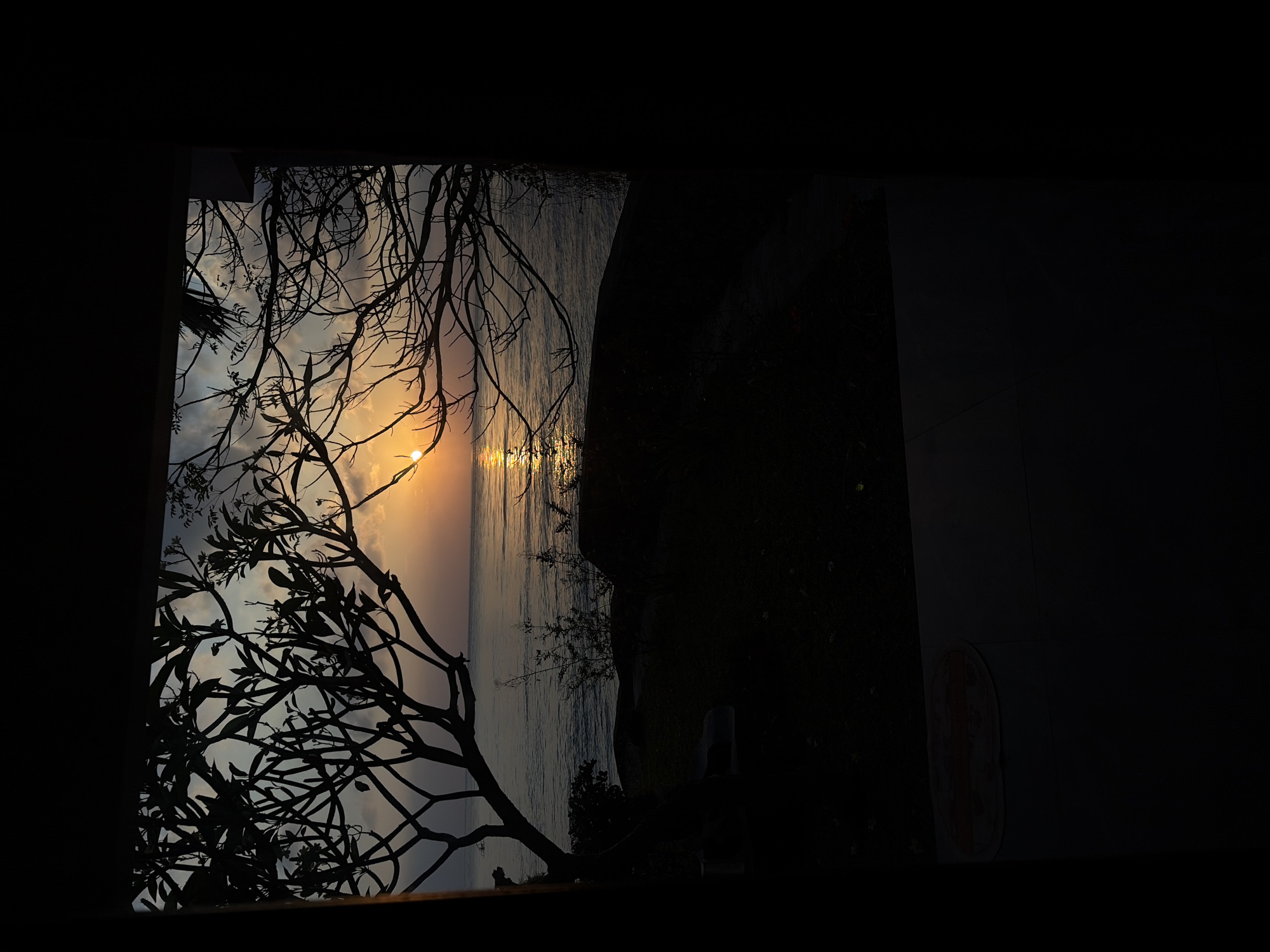 This image captures a silhouette of a tropical landscape at sunset, viewed through an opening that frames the scene. The sun is setting over a calm body of water, casting a golden glow on the surface. Silhouetted trees with thin branches frame the view on the left and right sides. On the left, a tree with fuller foliage is prominent. The ground in the foreground is dark and appears to be covered in grass and some rocks. The sky is partly cloudy with hues of orange and blue. The scene is located in Abang, Indonesia.