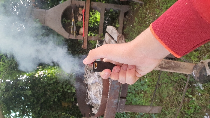 A person's right hand is holding a black lighter, the top of the lighter is slightly pressed down and smoke is emanating from the top of the lighter. The hand is wearing a red long-sleeved shirt. The person is standing next to a charcoal grill with a metal grate on the top, and there is ash and charred material on the grate. The grill is sitting on a wooden stand. In the background there is a brick structure, and the structure appears to have been constructed for barbecuing or grilling. There are a few items on the top shelf, such as a metal spatula, a pile of food and some leaves. The grill is located on a grassy, outdoor area and there is green vegetation and a few trees around. The location is in Libourne, France, at coordinates 44.907 latitude and -0.238 longitude.