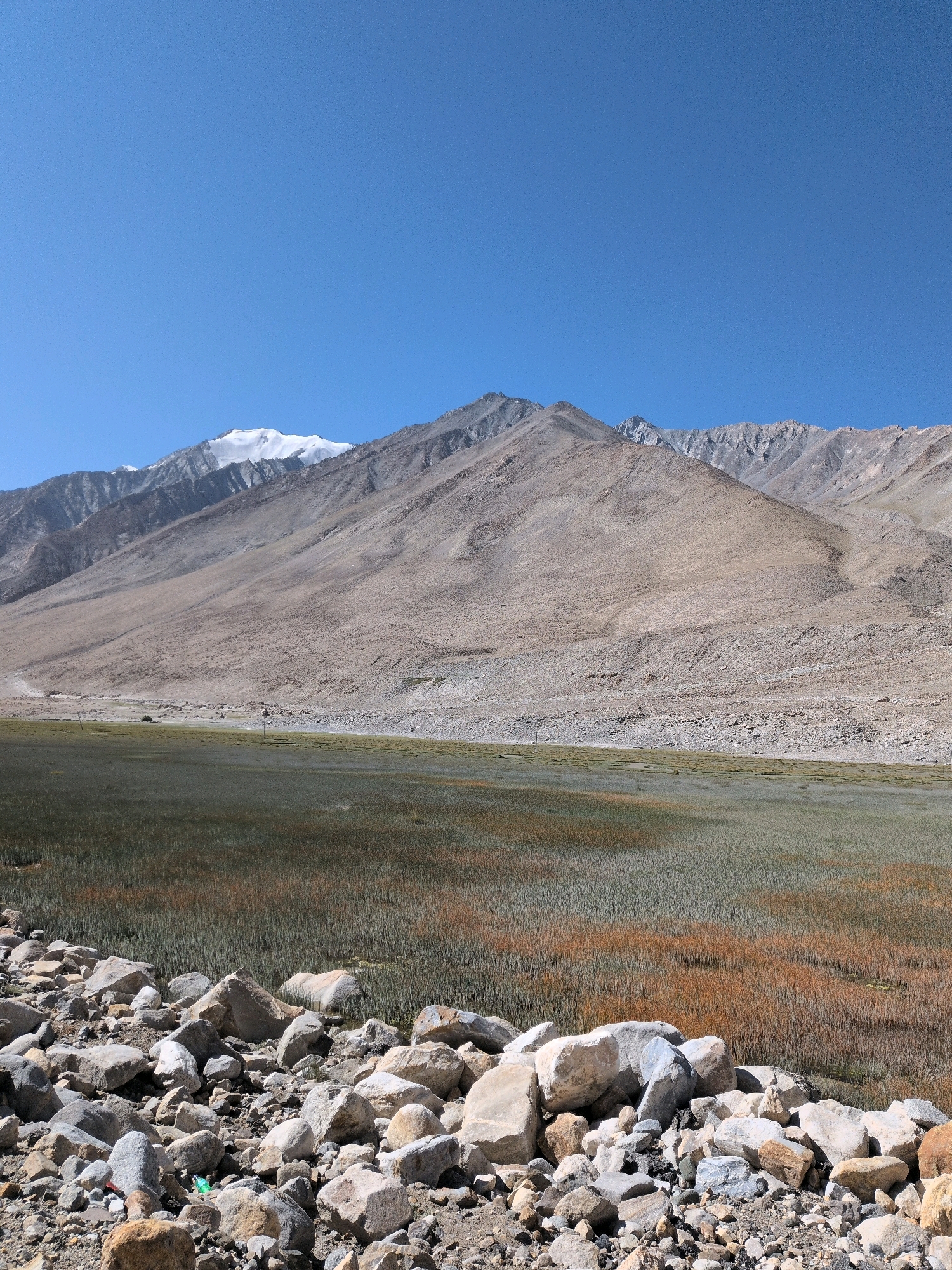 The image depicts a vast, arid mountain landscape under a clear blue sky. In the foreground, a pile of various-sized rocks and pebbles fills the lower portion of the frame. Beyond the rocks is a wide, marshy area with dry grass and some patches of green vegetation, suggesting a shallow body of water or damp ground. In the middle ground, a series of barren, sloping mountains stretches across the horizon. The mountains are predominantly light brown and rocky, with some areas of scree. The peaks of the furthest mountains in the background are capped with snow, indicating a high altitude. There are no people or man-made structures visible in the scene, aside from what might be a faint track or road on the far slope of the mountains. The overall impression is one of a remote and rugged natural environment in a dry, mountainous region.