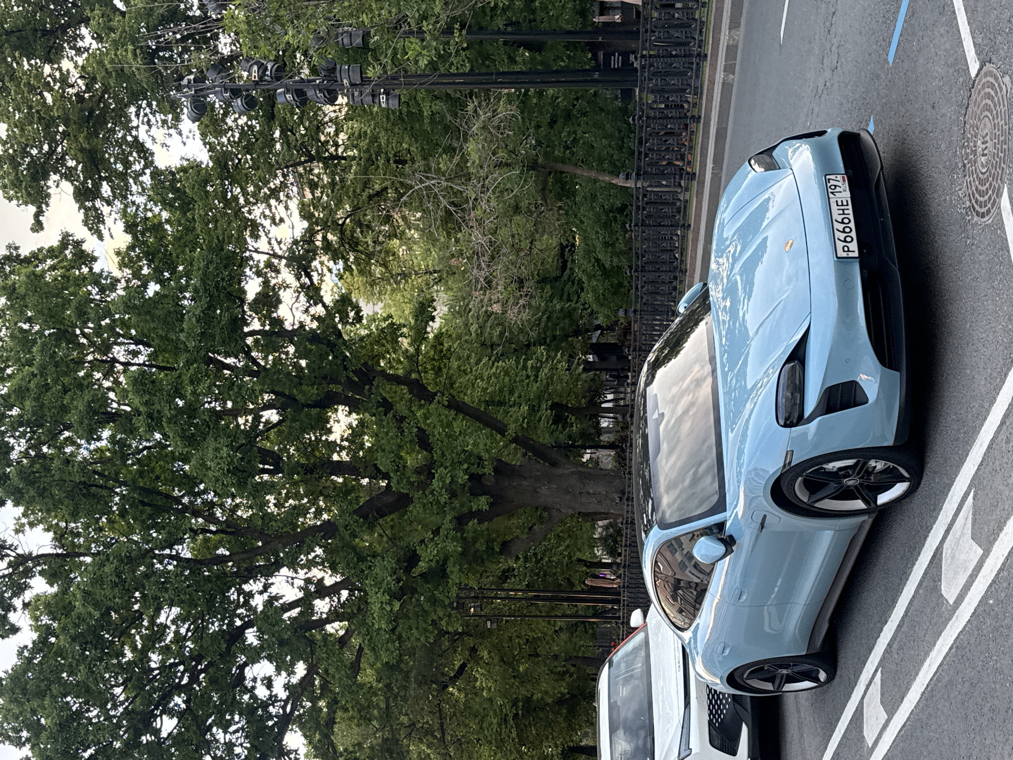 This media file captures an outdoor street scene, rotated 90 degrees clockwise, primarily featuring a light blue car parked alongside a street with trees in the background.
The central subject is a light blue, likely electric, car (resembling a Porsche Taycan) parked parallel to a white line on asphalt pavement. The car is seen from its front-left quarter, displaying sleek lines and black multi-spoke wheels. Its Russian license plate, clearly visible on the front bumper, reads "P666HE 197 RUS". Part of a white vehicle is visible behind the blue car.
To the left of the car, a lush canopy of green trees dominates the scene, suggesting a park or a tree-lined boulevard. A black metal fence runs along the edge of this green area. Among the trees, several black poles fitted with multiple light fixtures (resembling stage or floodlights) are visible, hinting at either permanent decorative lighting or setup for an event.
The pavement next to the car features white lane markings and a distinctive blue painted line. A circular manhole cover with concentric patterns and "KK" (or similar Cyrillic characters) etched on it is visible near the front of the car. The sky, partially visible through the tree canopy, appears bright, suggesting a daytime setting, possibly under overcast conditions.
No specific activity or people are depicted. The license plate's "197 RUS" code confirms the location as Moscow, Russia, aligning with the provided context.