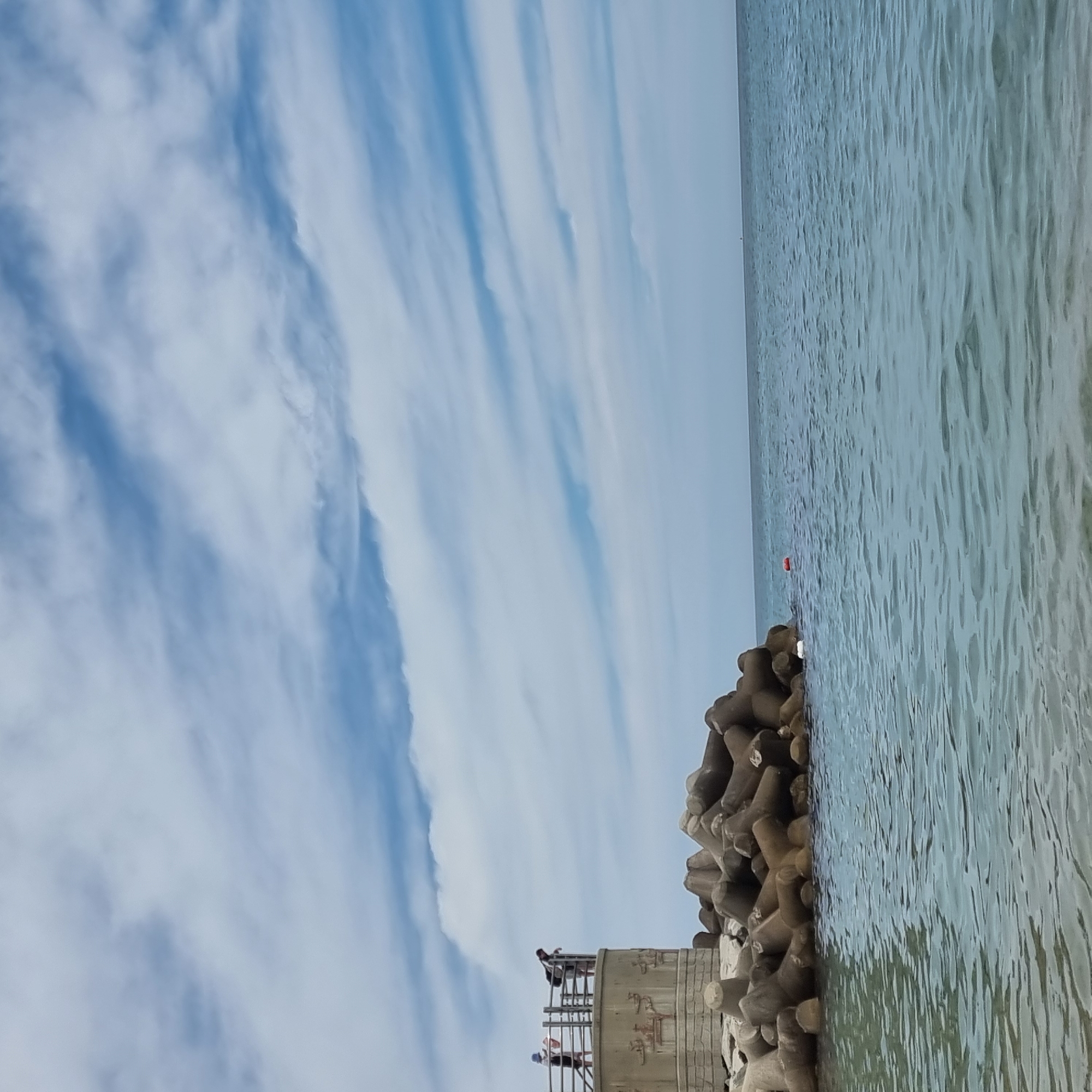 This is an image of the coastline in Gangneung-si, South Korea. In the foreground, the clear, blue ocean laps gently against the shore. In the midground, a concrete structure with railings stands on a breakwater, adorned with relief carvings. Two people are visible on top of this structure, one facing away from the camera, and another leaning over the railing, looking down. A pile of tetrapods, large concrete structures designed to protect coastlines from erosion, sits to the right of the concrete structure, partially submerged in the water. Further out in the ocean, a small red buoy is visible. The sky above is a mix of blue and white clouds, with a light breeze creating wispy patterns. The overall scene depicts a peaceful day at the beach, with a focus on the man-made elements of coastal defense and a natural, serene environment.