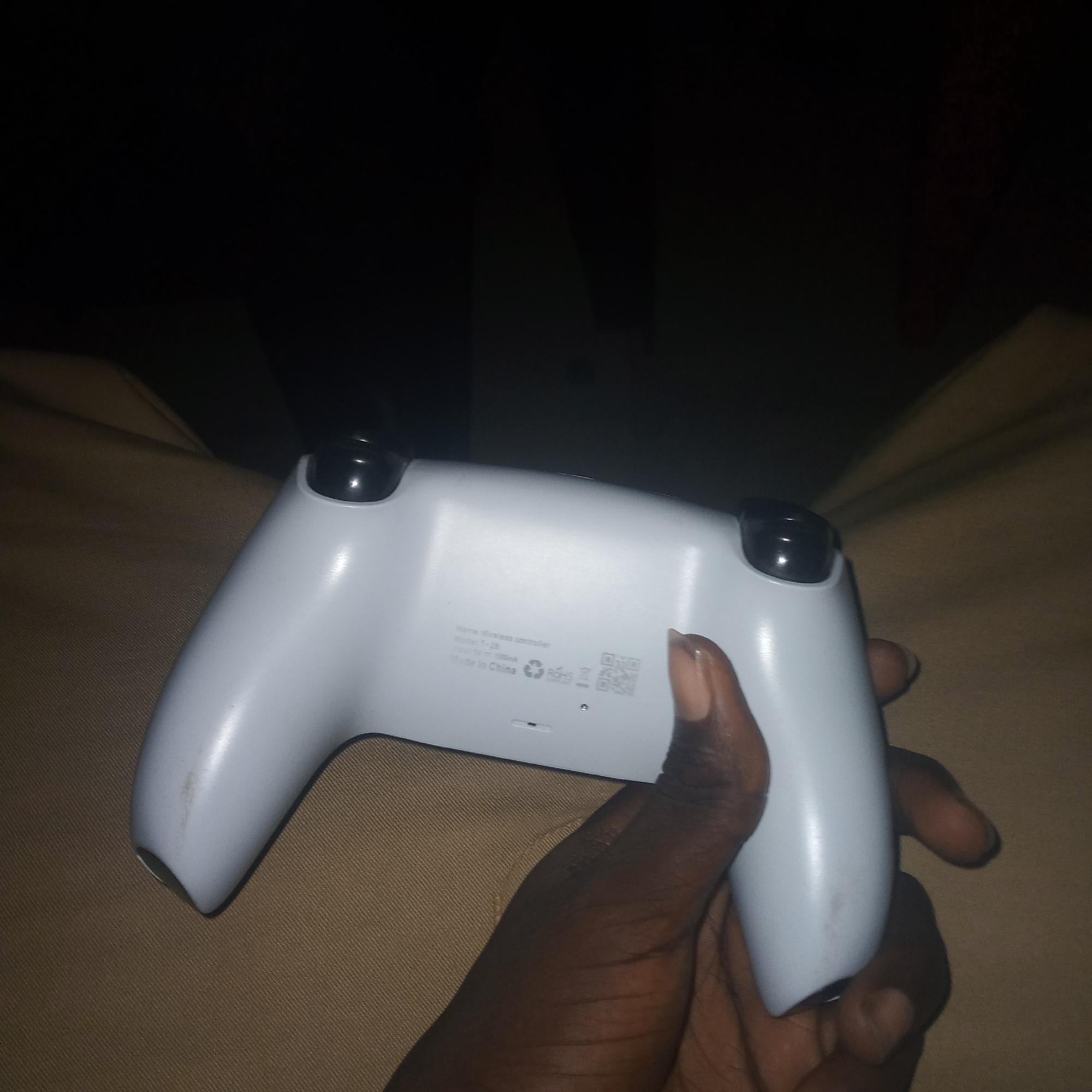 A hand is holding a white wireless controller against a tan, textured surface. The controller is oriented horizontally, with the analog sticks visible at the top. There is some text and a QR code on the back of the controller. The image is dark, with the primary light source appearing to come from above and slightly to the right. The background is mostly black and out of focus.
