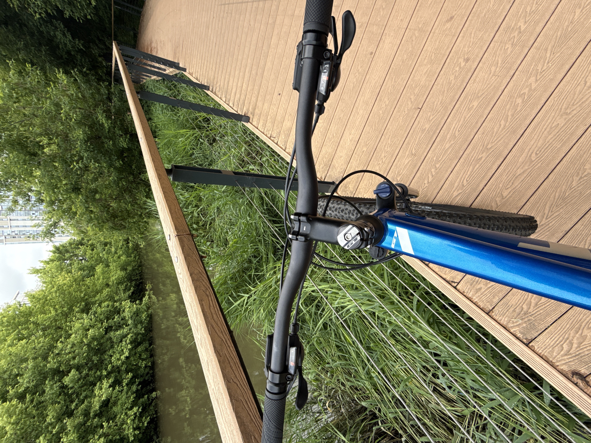 A first-person perspective shot shows a blue bicycle's handlebars on a wooden boardwalk next to a tranquil body of water. The boardwalk curves to the right, bordered by a railing composed of wooden beams and metal posts with thin cable railings. Lush green trees and foliage line the opposite bank of the water, with a tall building visible in the distance. The bicycle has a black grip, gear shifters, and brake levers on the handlebars. The scene is set in a park in Moscow, Russia.