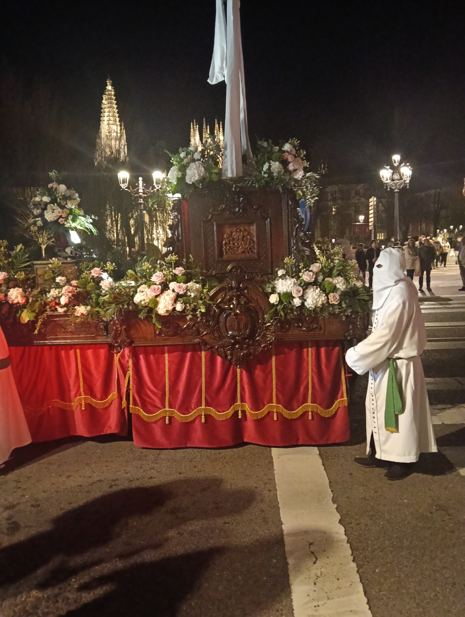 At night, an image captures a religious procession in Burgos, Spain. The scene is illuminated by streetlights and the faint glow from a building in the background. In the foreground, a dark wooden structure adorned with an array of flowers and draped with a red cloth featuring gold tassels is being paraded. Standing alongside the structure is a person clad in a white robe with a pointed hood, indicative of religious participation. The person wears a green sash. They are standing on a zebra crossing painted on the asphalt street. In the background, several people are seen, indistinctly lit, watching the procession. The architecture suggests the setting is a European city.