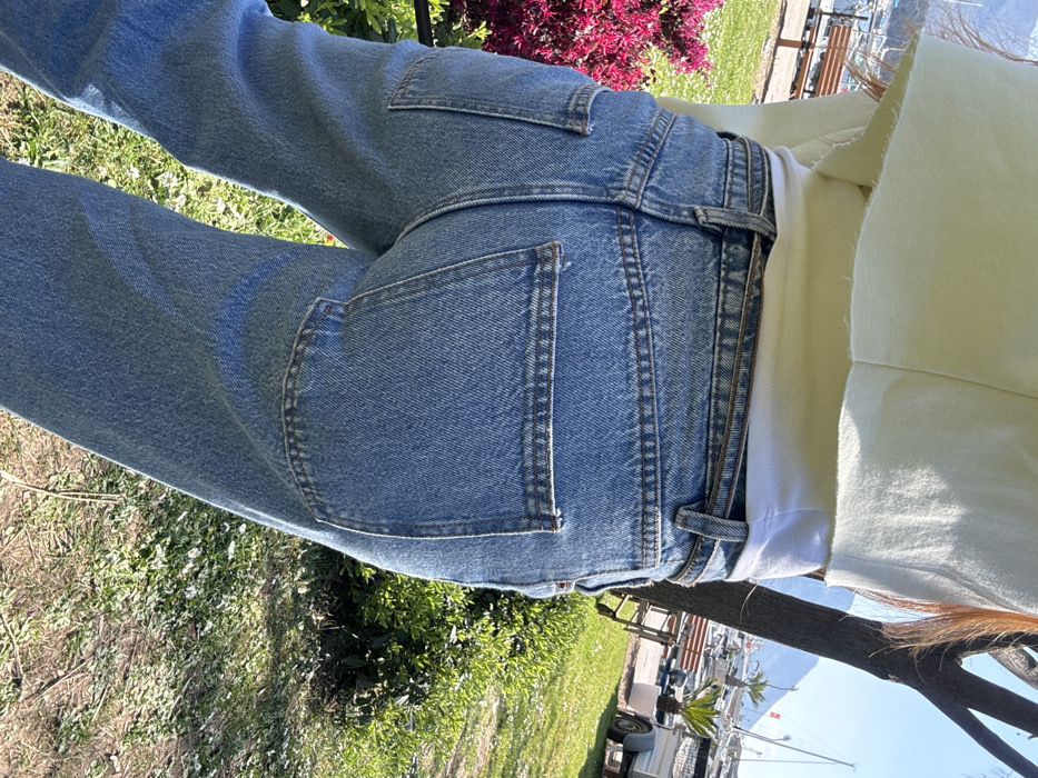 The image displays the back of an individual from the waist down, standing in a sunny outdoor park area adjacent to a marina in Budva, Montenegro. The person is wearing light blue denim jeans with prominent stitching and rectangular rear patch pockets, a white undershirt, and a light yellow, raw-hemmed cropped top. Long, reddish-brown hair is visible over the left shoulder. To the left, a bush with vibrant pink flowers is present. The background features a harbor with several sailboats moored, visible beyond a paved path or walkway. A light brown wooden bench is situated near the marina. On the right side of the frame, green shrubs, a tree trunk, a white concrete planter containing a small palm tree, and a small utility trailer are visible. Distant mountains rise under a clear blue sky in the far background.