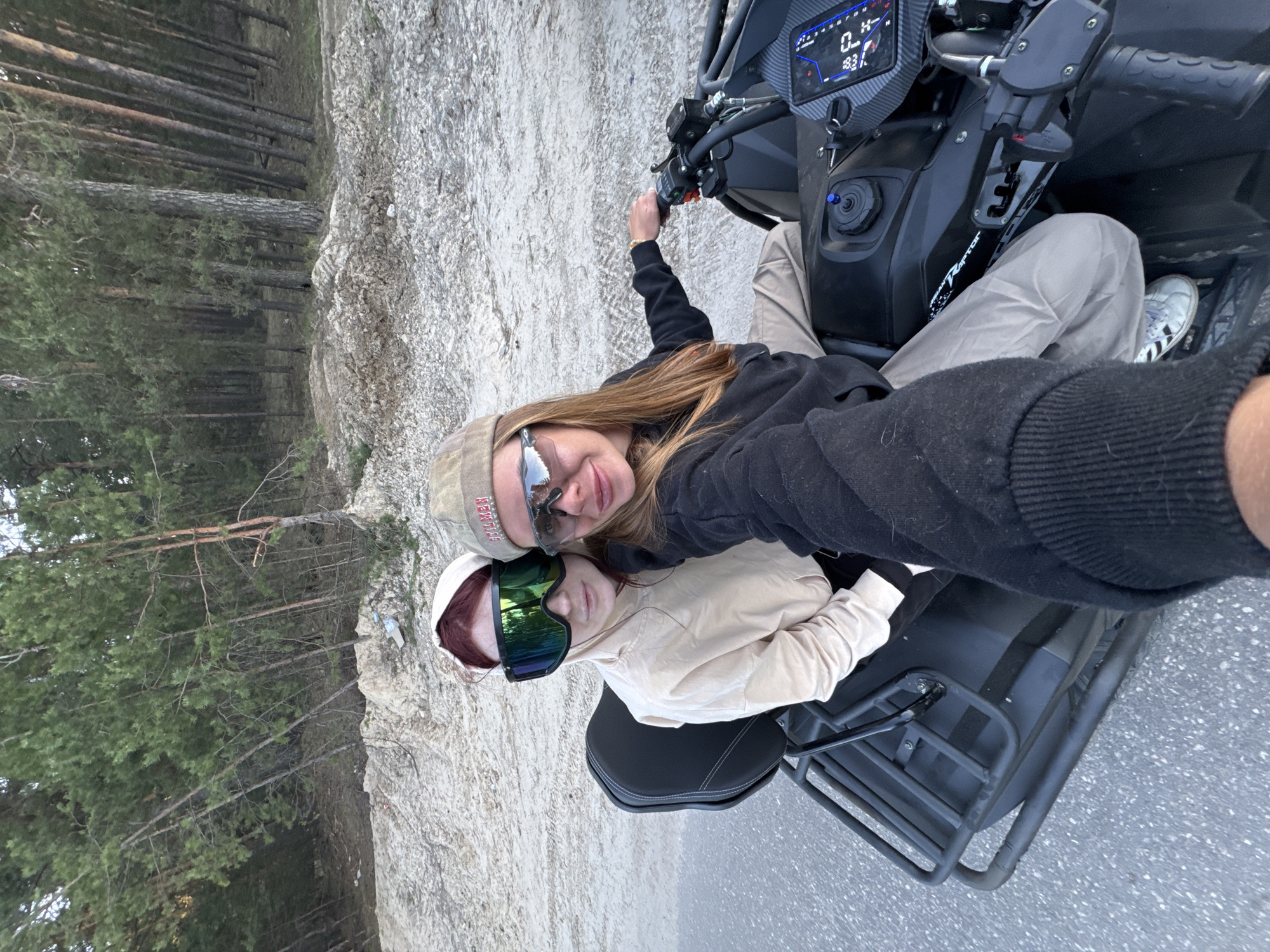 Two individuals are captured in a selfie while riding an ATV. One person is wearing a baseball cap and sunglasses, while the other is wearing goggles. They are positioned on the ATV in front of a backdrop of a forest and what appears to be a sandy or rocky embankment. The location is Bol'shoye Kozino, Russia.