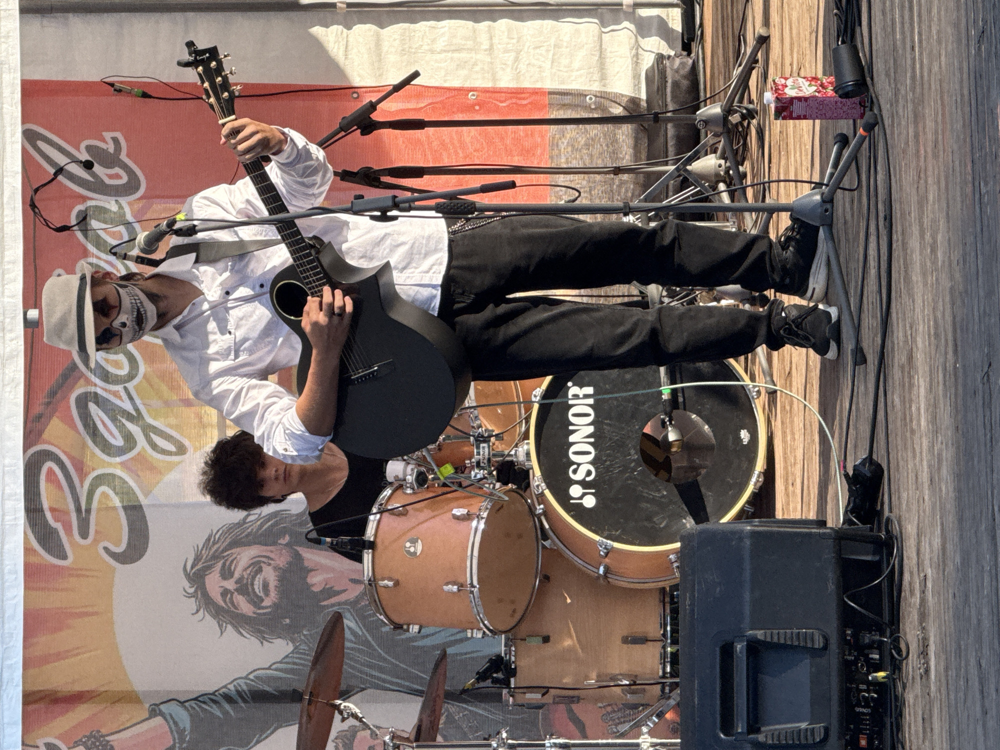 A male musician wearing a white shirt, black pants, a light-colored fedora, and skull makeup is playing an acoustic guitar on a wooden stage. He is performing in front of a banner with a graphic of a cheering man and Cyrillic text that reads "Зотов". To his left, a drummer with dark, curly hair is seated behind a drum set. The stage has audio equipment, including a speaker and microphones. The scene appears to be an outdoor music event.