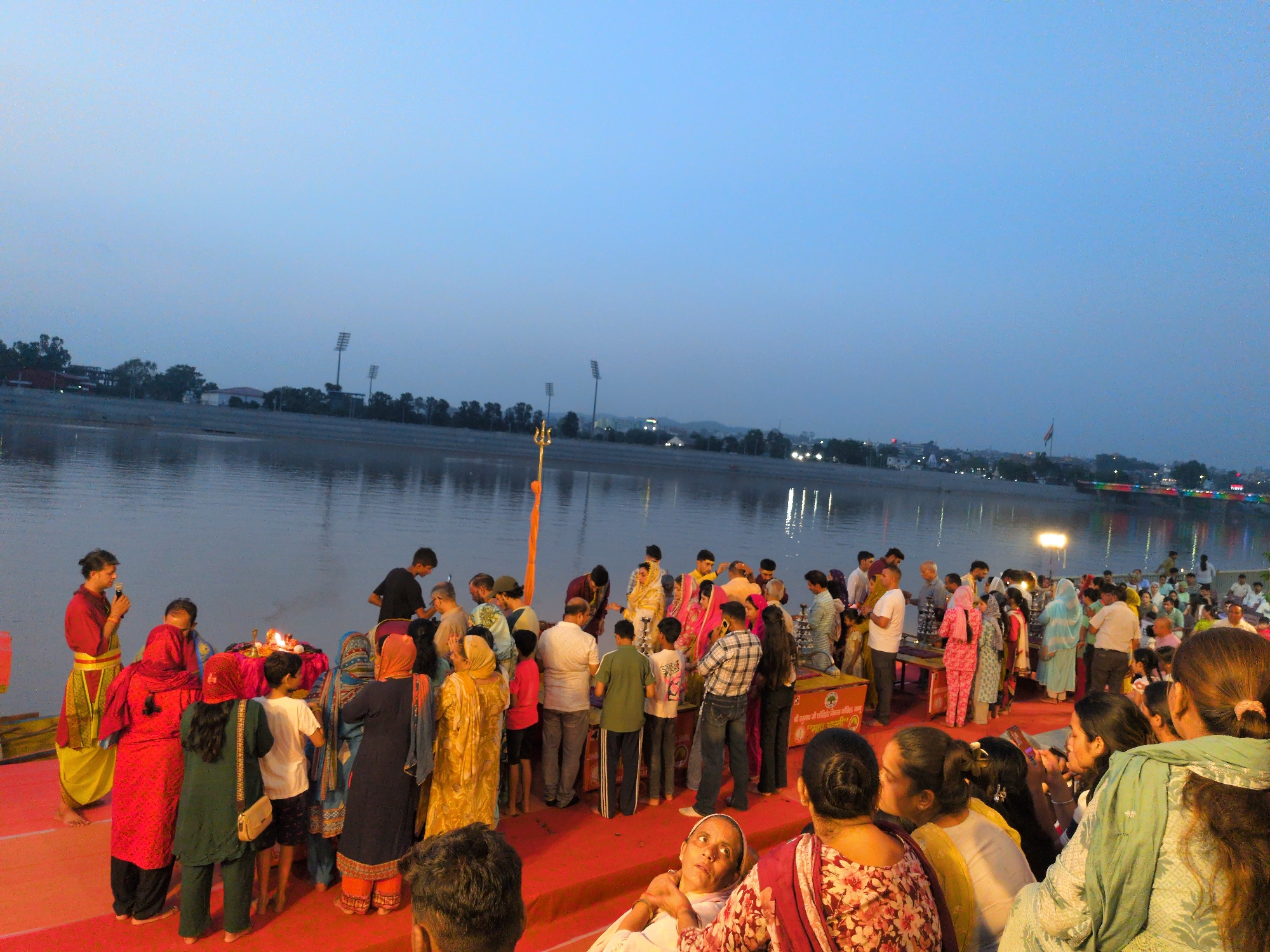 This is a wide-angle, eye-level shot of a gathering of people on a red carpeted platform near a body of water, possibly the Tawi River, in Jammu, India. The sky is a clear, pale blue, indicating it is either dawn or dusk. In the background, across the water, lights from buildings and streetlights are visible, along with some trees and what appear to be stadium lights.
The foreground is dominated by a large crowd of people, many dressed in traditional Indian attire, including saris and kurtas. Some individuals are standing, while others are seated or kneeling. A few people are gathered around a small altar or table where a fire is burning, suggesting a religious ceremony or ritual is taking place. There are multiple individuals holding what appear to be mobile phones, some of whom are taking pictures or videos. Several people are interacting with each other, with some looking towards the altar and others looking towards the camera or elsewhere. There is a prominent trident-like object with a flag standing in the water near the bank. The overall atmosphere appears to be one of communal gathering and participation in a cultural or religious event.