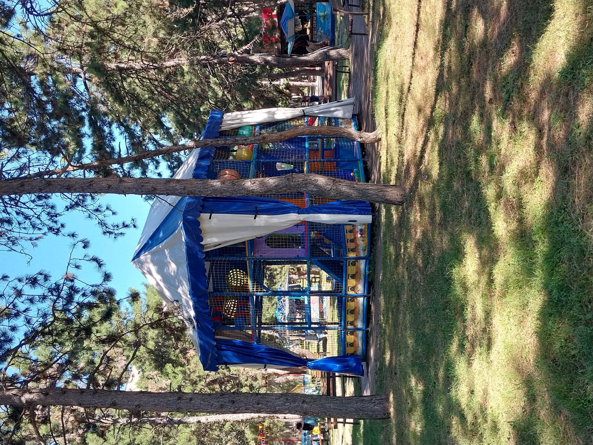 The image captures an outdoor children's playground structure in Dnipro, Ukraine, under a clear blue sky on a sunny day. The playground is a multi-level, enclosed structure with a blue and white tent-like canopy. It features blue netting walls, colorful climbing elements, slides, and balls visible within the play area. Blue drapes hang from the sides of the canopy. The structure is situated on a grassy area with several mature trees providing shade. In the background, other playground equipment, benches, and a kiosk or small building can be seen. A few people are present in the distance, one standing near a bench and another near the kiosk. The overall scene depicts a recreational area in a park-like setting.