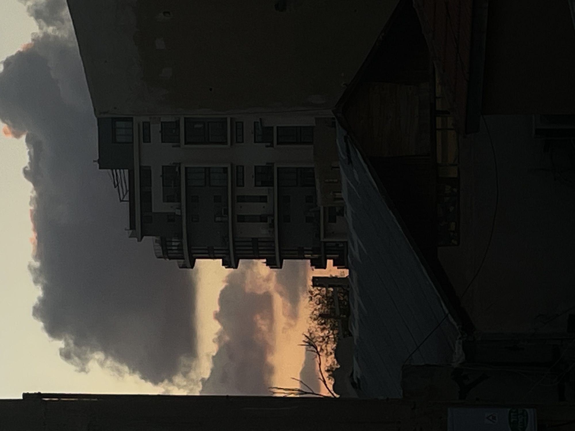 The image depicts an urban scene in Tel-Aviv, Israel, presenting a multi-story residential building under a cloudy sky, appearing to be at sunset, with shades of orange and gray. The building is characterized by balconies and multiple windows. The foreground includes portions of adjacent buildings or structures, creating a framed perspective. Details are obscured by shadows.