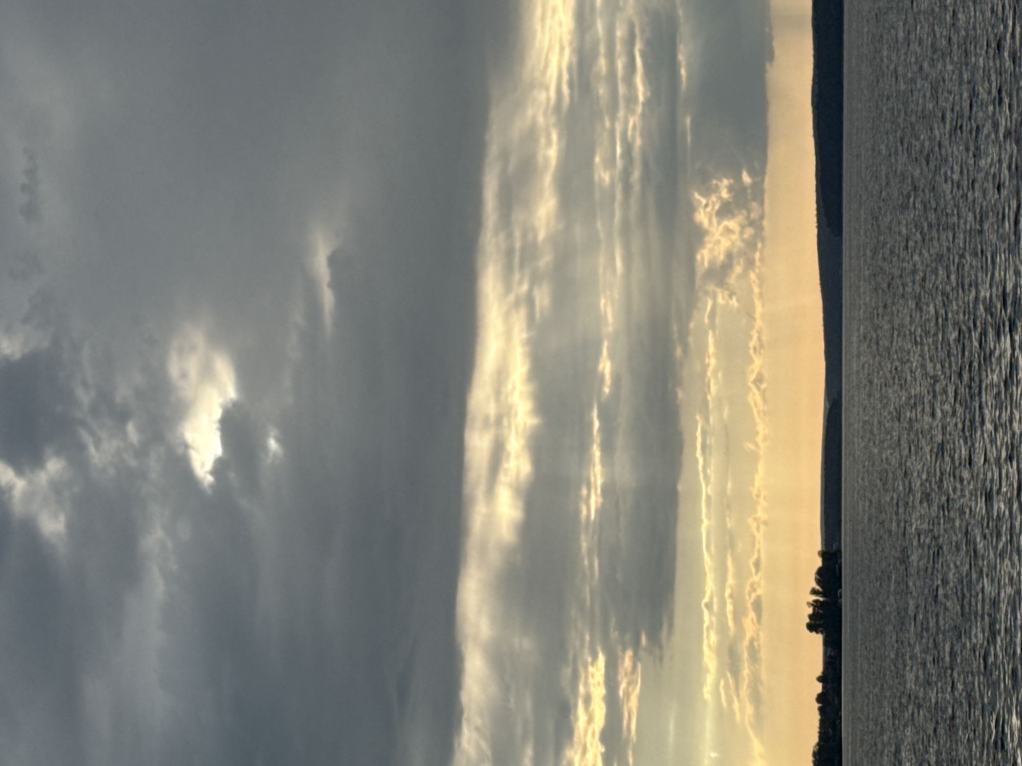 The image depicts a panoramic view of the Kama River in Votkinsk, Russia, under a dramatic sky. Dark, heavy clouds dominate the upper portion of the frame, with shafts of light breaking through, creating a crepuscular effect. The lower half of the image is occupied by the calm, rippling surface of the river, reflecting the subdued light. In the distance, a dark, tree-lined shoreline is visible, punctuated by a cluster of trees on a small peninsula. The overall mood is somber and atmospheric, characteristic of a moody late afternoon or early evening. There are no people, animals, or man-made structures, besides a faint suggestion of buildings on the far shore, visible on the left side of the frame.