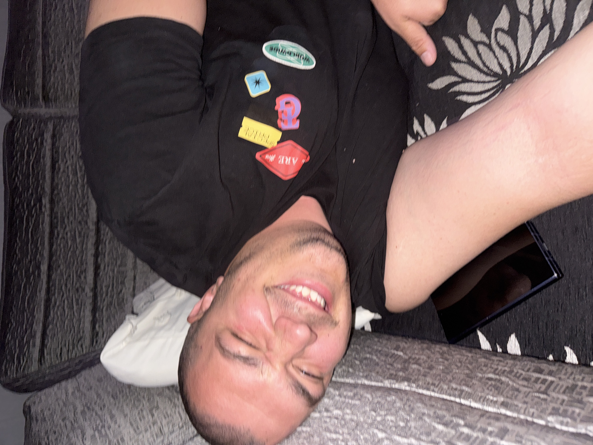 A person is lying on a gray couch with a white pillow. The person is wearing a black t-shirt with patches. A black tablet is next to the person on a decorative pillow. The location is not identifiable as Bozhurishte, Bulgaria.