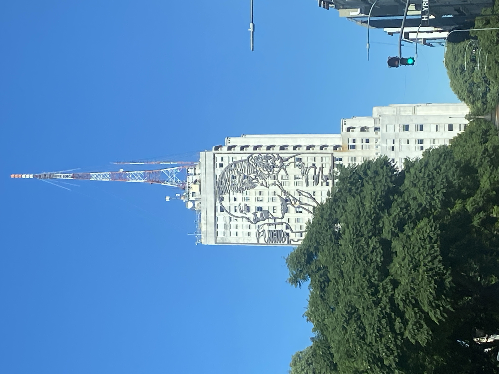 The image presents a vibrant urban scene under a clear, bright blue sky, captured during what appears to be a sunny day, likely midday or early afternoon.
In the mid-ground, a large, light-colored building, possibly a high-rise office or apartment block, stands prominently. Its facade is adorned with a distinctive, large-scale artwork or mural executed in dark lines, depicting an abstract or skeletal human form, possibly a head or skull, intricately integrated across multiple floors. The building features numerous windows, some with visible shutters or awnings.
To the left of this building, a very tall communication or broadcast tower, constructed with red and white lattice-work, stretches into the sky, equipped with various antennas and transmission gear. In the foreground and mid-ground on the right side of the image, a dense mass of rich green foliage from large trees partially obscures the lower parts of the building.
Above the tree line on the far right, urban infrastructure elements are visible: a traffic light displaying a green signal, and a partial sign or text on a building reading "H. YRI". This likely refers to "Hipólito Yrigoyen," a known street or location in Buenos Aires, Argentina, aligning with the provided context.
The scene is static, devoid of people or specific activities, offering an observational snapshot of an architectural and artistic urban landscape.