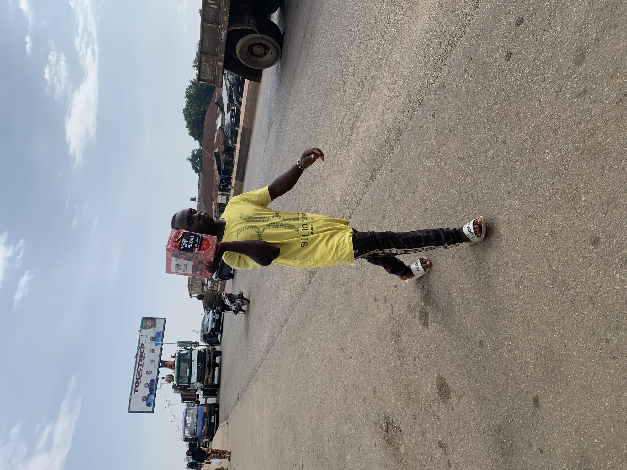 A young Black man walks along a paved road in Jos, Nigeria, carrying a red and white box of Runall car batteries on his right shoulder. He is wearing a yellow t-shirt with a graphic design and the word "BILLIONAIRE" printed on it, and dark pants with red and white stripes down the sides. He has white slides on his feet with "LUOFU" printed on them. The man is walking towards the right side of the frame. In the background, several vehicles are visible, including a blue truck, a large green truck, black cars, and a motorcycle. A billboard displaying a political message with the word "TOGETHER" is visible in the distance. The road appears to be a major thoroughfare with buildings and trees lining the side. The sky is overcast with scattered clouds.