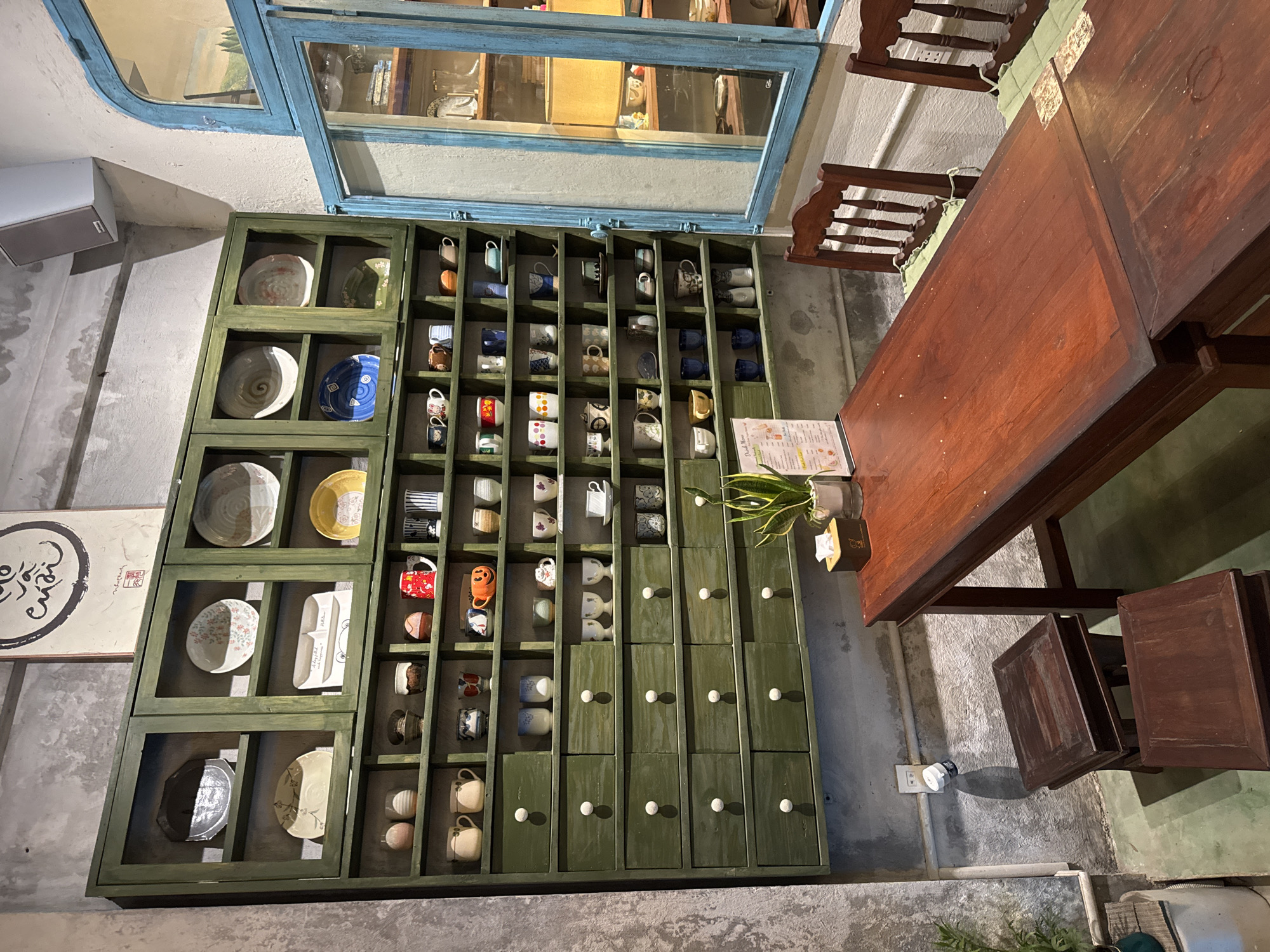 The image shows an indoor scene in what appears to be a cafe or restaurant. A large, dark wooden table dominates the right side of the frame, with matching chairs positioned around it. A decorative cabinet is visible along the wall, featuring shelves filled with various ceramic dishes, mugs, and what appear to be decorative boxes at the bottom, each having a white knob. A blue-framed mirror hangs above the cabinet, reflecting a glimpse of the interior. To the left of the cabinet, there is a framed print with Asian calligraphy. The lighting is somewhat dim, creating shadows and highlighting the textures of the wood and concrete walls. The scene suggests a stylized, possibly vintage or rustic aesthetic. It is difficult to confirm the location in Đà Nẵng, Vietnam, without further information.
