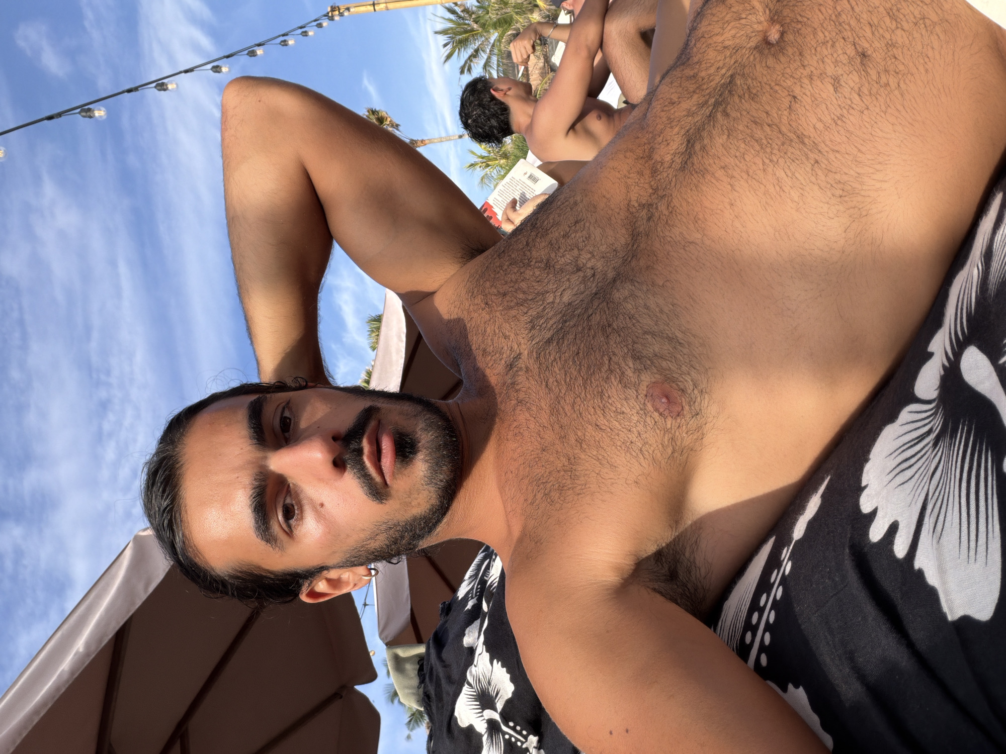 The image shows a man with dark hair, a beard, and mustache, lying on a lounge chair under a beach umbrella. He is wearing a black and white patterned fabric. His right arm is bent behind his head, and he is looking at the camera. In the background, another man is sitting and reading a book, with palm trees and a clear blue sky with clouds visible. There are also string lights overhead, suggesting an outdoor setting like a resort or beach club in North Kuta, Indonesia. The lighting indicates it is daytime.