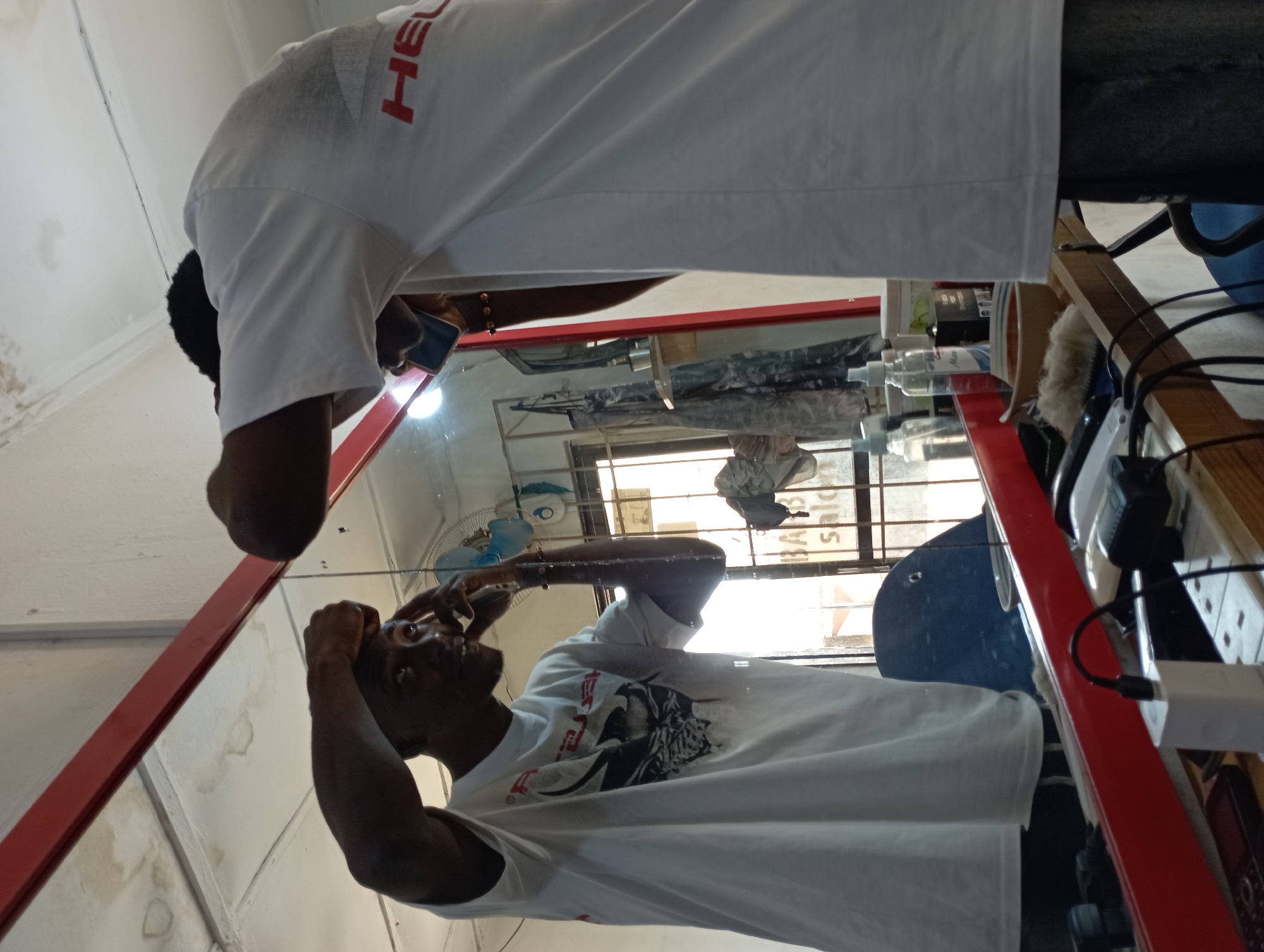 The image portrays an indoor scene, likely a barbershop or salon, featuring a man and his reflection in a large, rectangular mirror framed in red. The walls are light-colored, with some visible stains or marks.
A dark-skinned man with short, dark hair is leaning forward, looking into the mirror. He is wearing a white short-sleeved t-shirt with red text, partially visible as "HEAD," on the back shoulder. His reflection in the mirror shows him looking slightly upwards, with his hands positioned near his head, suggesting he is grooming or examining his appearance. In the reflection, his t-shirt displays a black graphic print of a face or figure, and he wears a dark beaded bracelet on his right wrist.
The mirror's surface reflects several items arranged on a counter or shelf, including multiple transparent bottles, electronic chargers, and cables. A white oscillating fan is also visible in the reflection. Through a window reflected in the mirror, bright daylight is evident, indicating it is daytime. The reversed text "BARBER SALON" is clearly legible on the reflected window, confirming the type of establishment. The scene suggests a functional, everyday setting within a commercial grooming space in Jos, Nigeria.