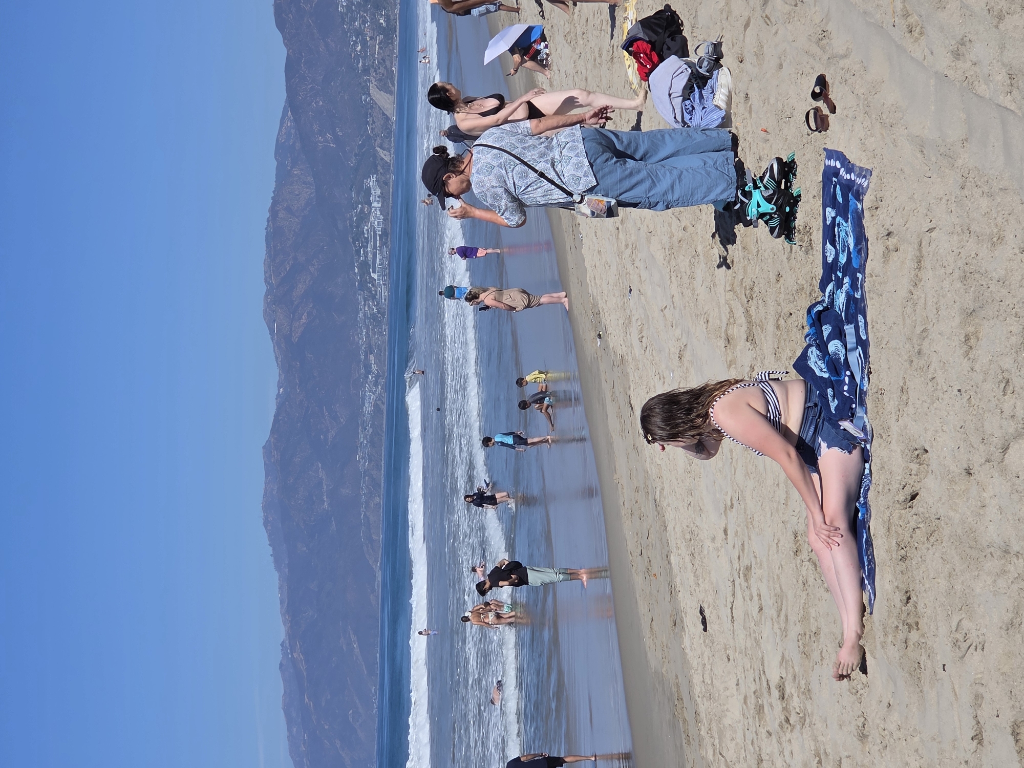 A panoramic, eye-level, full shot captures a sunny day at Santa Monica State Beach in Santa Monica, California. The ocean waves are gently rolling onto the shore, and in the distance, mountains form the backdrop under a clear blue sky. Several people are scattered across the beach and in the shallow water.
In the foreground, a woman with long, wet, brown hair sits on a blue and white patterned towel, facing left and shielding her eyes from the sun with her hand. She is wearing a black and white striped bikini top and denim shorts. To her right, a pair of black flip-flops rests on the sand, near a pair of rollerblades.
Further back, a woman wearing a patterned shirt, blue pants, and a black cap is drinking from a bottle. To her right, a woman in a black bikini stands with her arms at her sides. A white parasol is open and angled towards the ground behind her.
In the middle ground, people are wading in the ocean. A man in a black t-shirt and light blue pants stands in the water, facing away from the camera. To his left, a group of people, including children, are enjoying the water. Several other individuals are visible further out in the ocean and along the shoreline.
The sand is light brown and textured, with footprints and small indentations. Beachgoers' belongings, including clothing and bags, are visible near the woman in the foreground. The overall scene depicts a typical summer day at a popular beach destination.