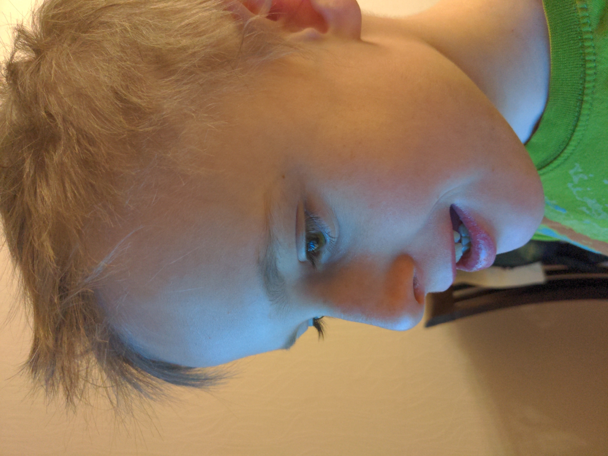 A close-up, slightly angled view of a child's face is presented, with the head tilted towards the right. The child has short, blonde hair and light skin. One eye is visible, along with the nose and partially open mouth, revealing teeth. The child is wearing a green shirt. The background is a light, neutral color. There is no indication of the location being in Moscow, Russia.