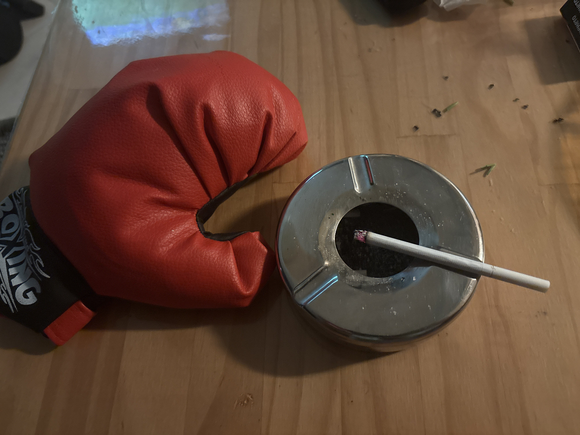 A red boxing glove rests on a wooden surface next to a metal ashtray. A lit cigarette rests in the ashtray, its cherry glowing. The scene is illuminated by overhead lighting, casting shadows on the table. Small dark specks, possibly debris, are scattered on the surface. The boxing glove has black and white text on its cuff, which appears to read "BOXING". The image is a close-up shot, focusing on the juxtaposition of the boxing glove and the ashtray with the lit cigarette. The overall impression is one of a momentary pause or a moment of contemplation. There are no people visible in the frame. The setting appears to be indoors.